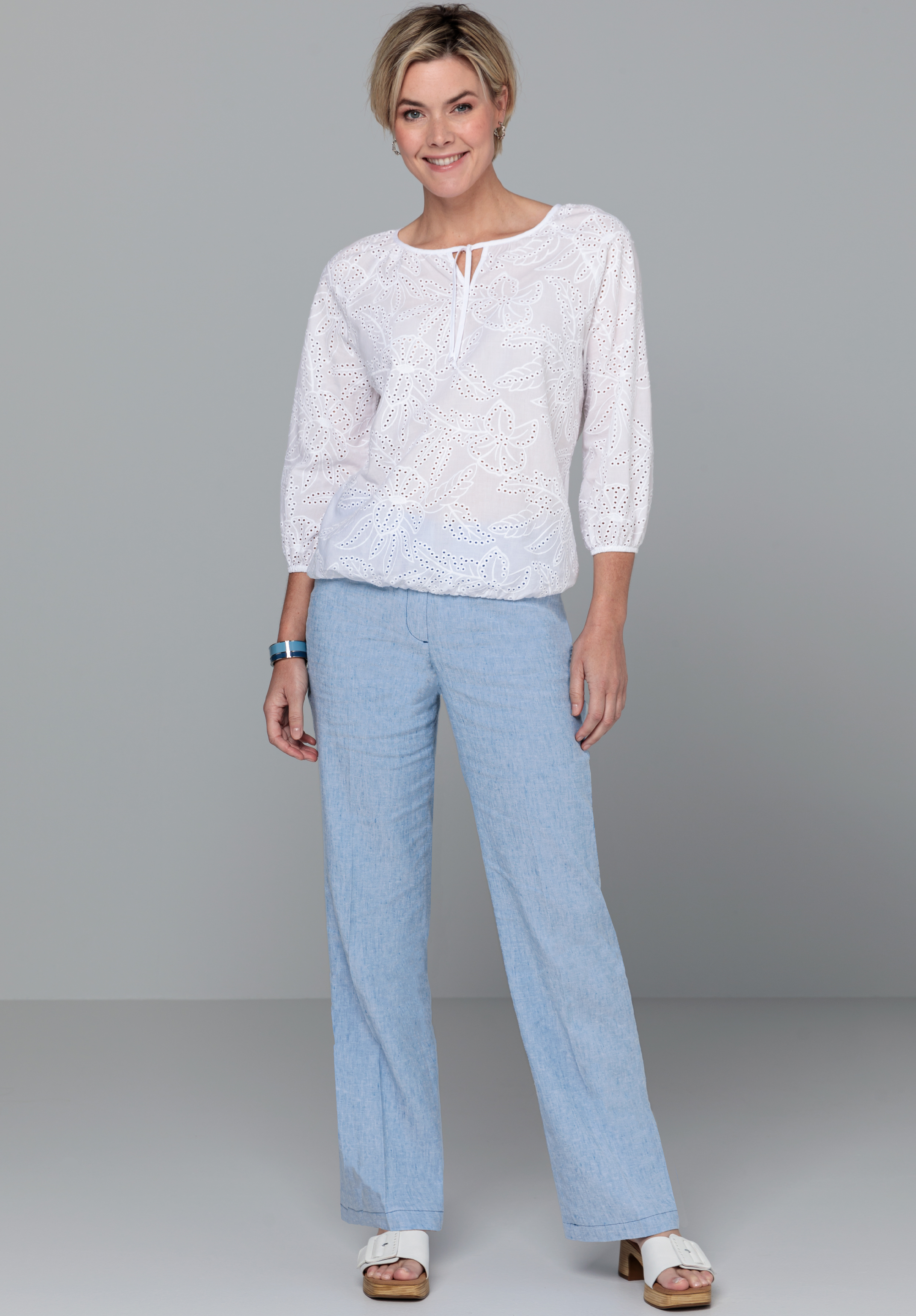 Eine Frau steht lächelnd vor einem schlichten grauen Hintergrund. Sie trägt eine weiße bestickte Bluse mit Dreiviertelärmeln, hellblaue Hosen mit geradem Bein, weiße Sandalen und eine blaue Armbanduhr.