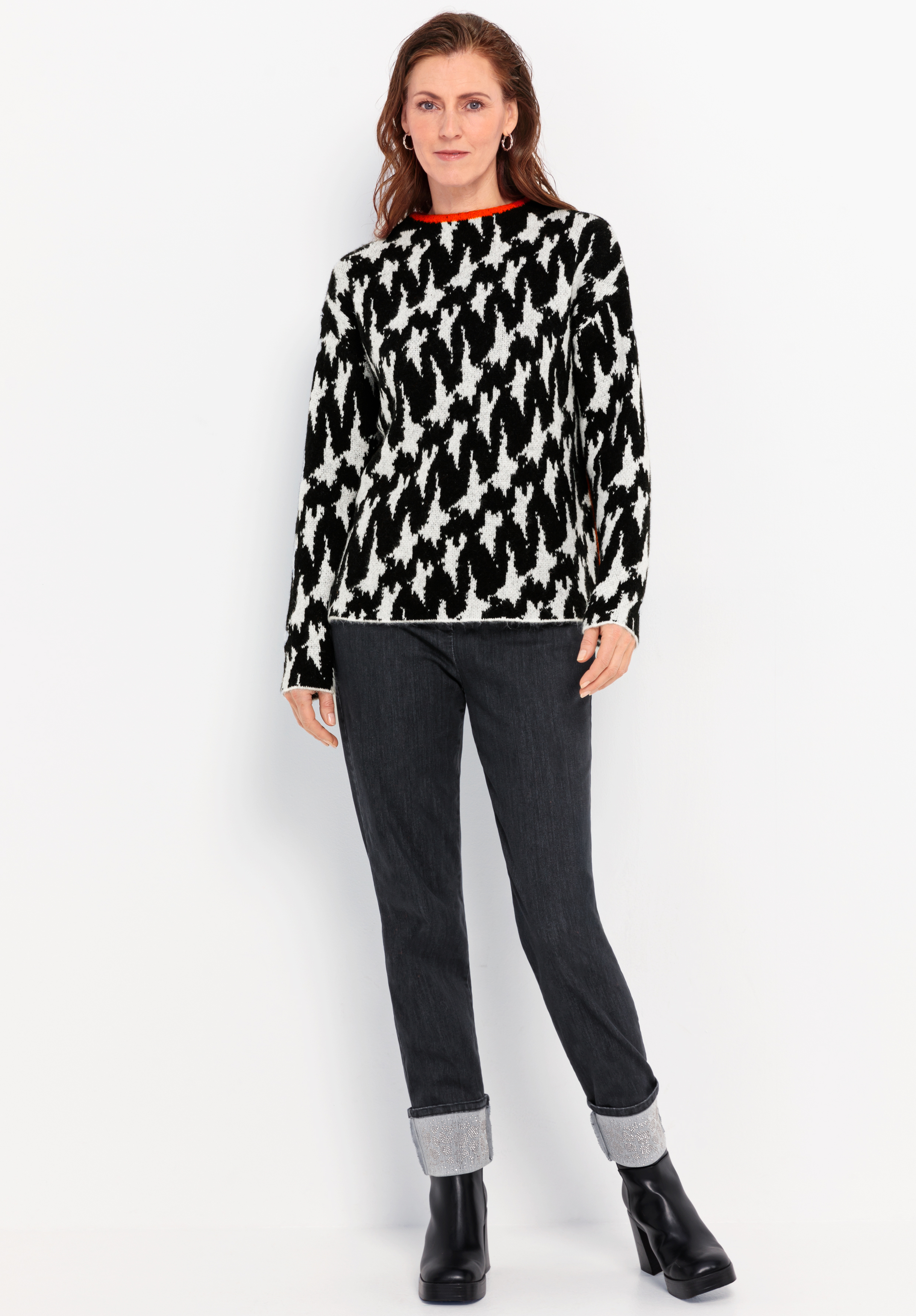 Vor einem weißen Hintergrund steht eine Frau, die einen schwarz-weißen Pullover mit Hahnentrittmuster, eine dunkle Jeans mit Umschlag und schwarze Stiefeletten trägt. Sie hat langes braunes Haar und einen entspannten Gesichtsausdruck.