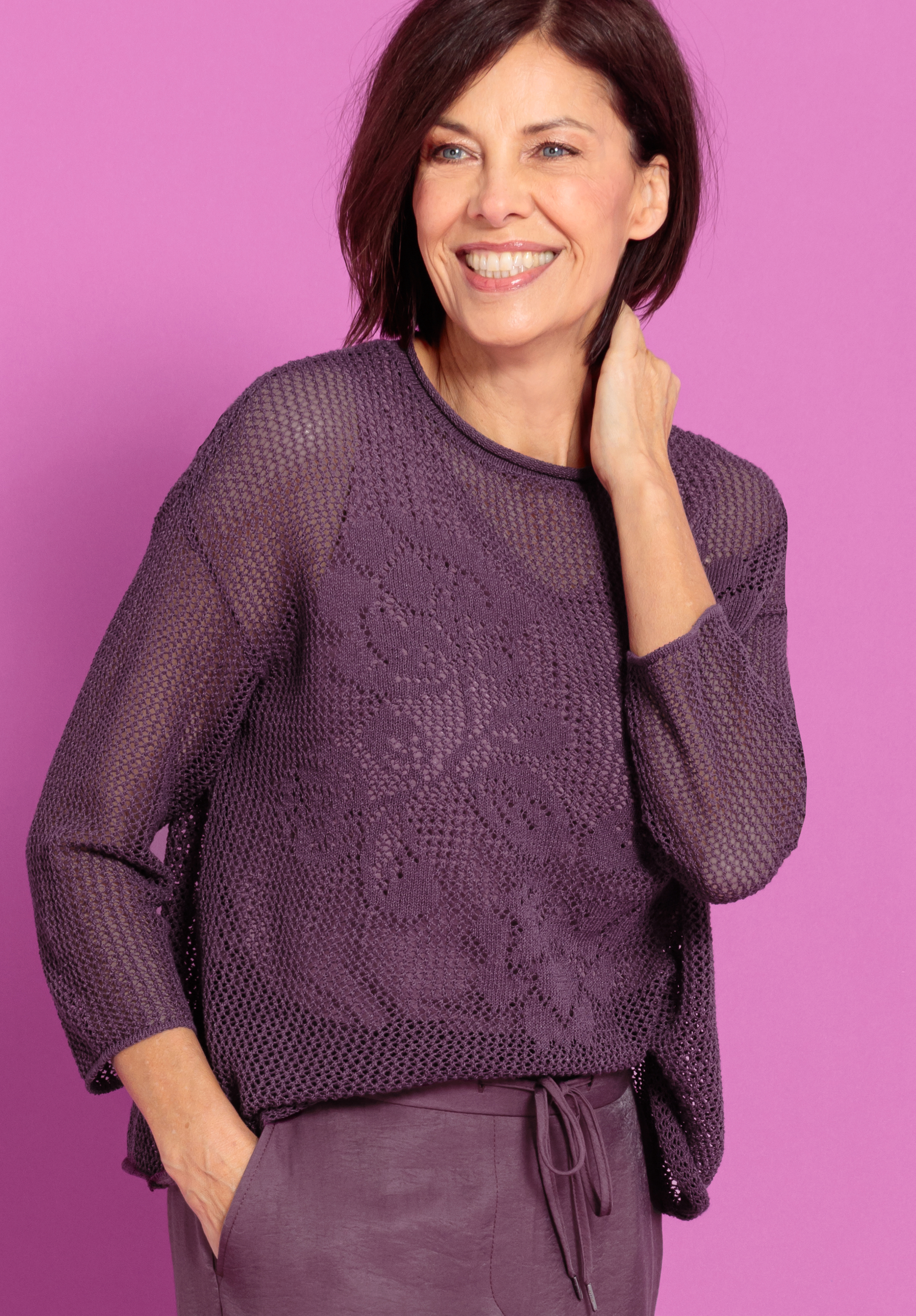 Eine Frau mit schulterlangen braunen Haaren, die einen lilafarbenen Strickpullover und eine dazu passende Hose trägt, lächelt, während sie vor einem rosa Hintergrund steht. Eine Hand steckt in ihrer Tasche, die andere berührt ihren Hals.