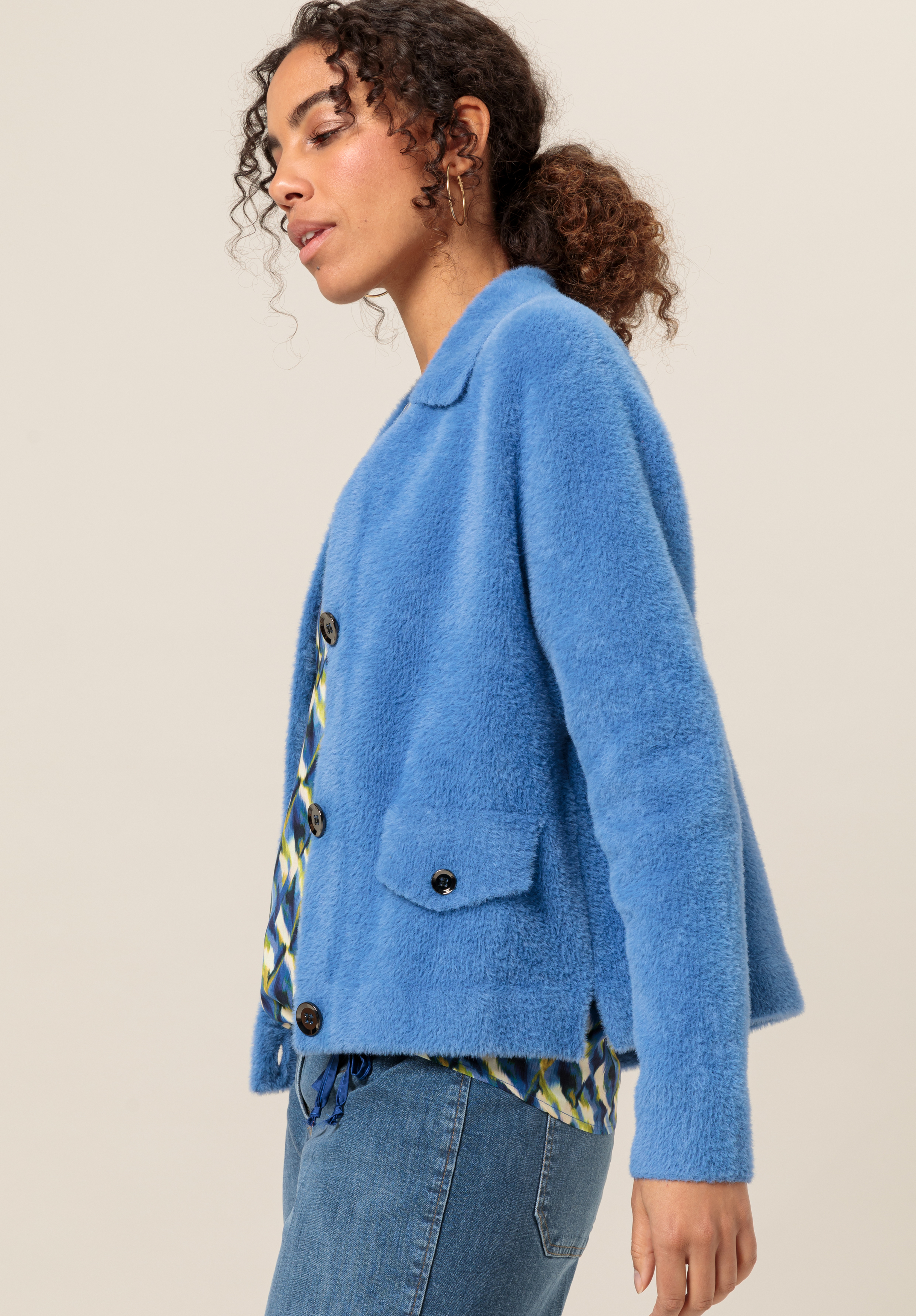 Eine Frau mit zurückgebundenem lockigem Haar trägt eine strukturierte blaue Jacke mit schwarzen Knöpfen und Vordertaschen über einem gemusterten Hemd und blauen Jeans und steht vor einem schlichten beigen Hintergrund.