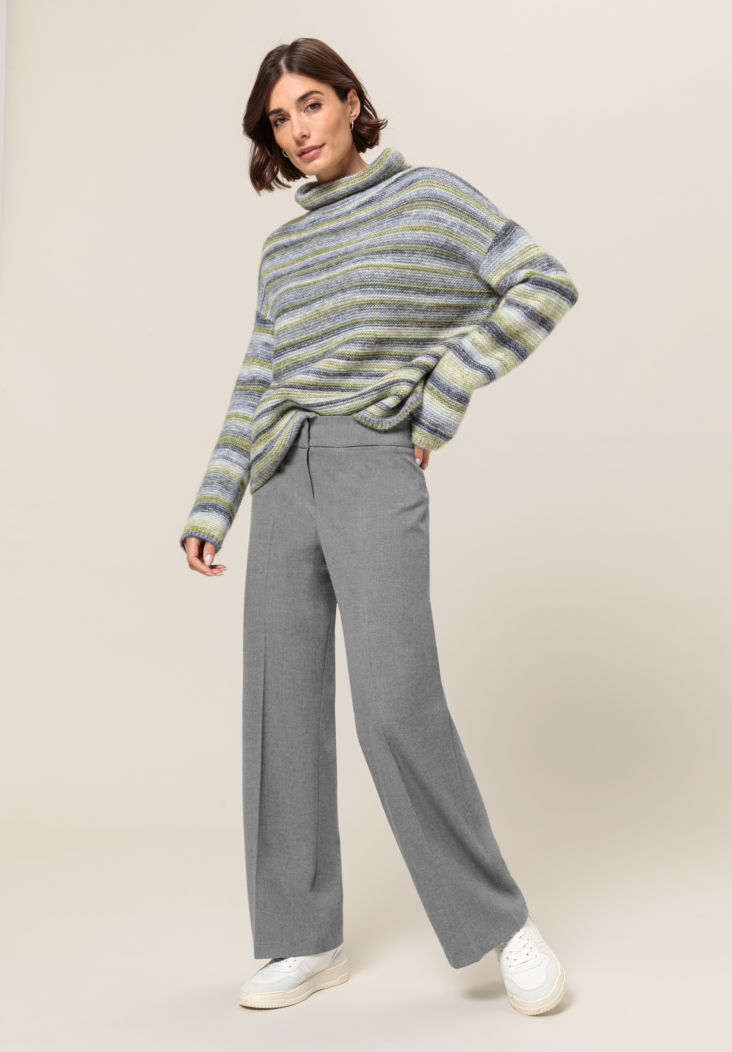 Eine Frau steht vor einem schlichten Hintergrund. Sie trägt einen lockeren, gestreiften Rollkragenpullover in Grau-, Grün- und Blautönen, eine weite graue Hose und weiße Turnschuhe. Sie hat kurze braune Haare und eine entspannte Pose.