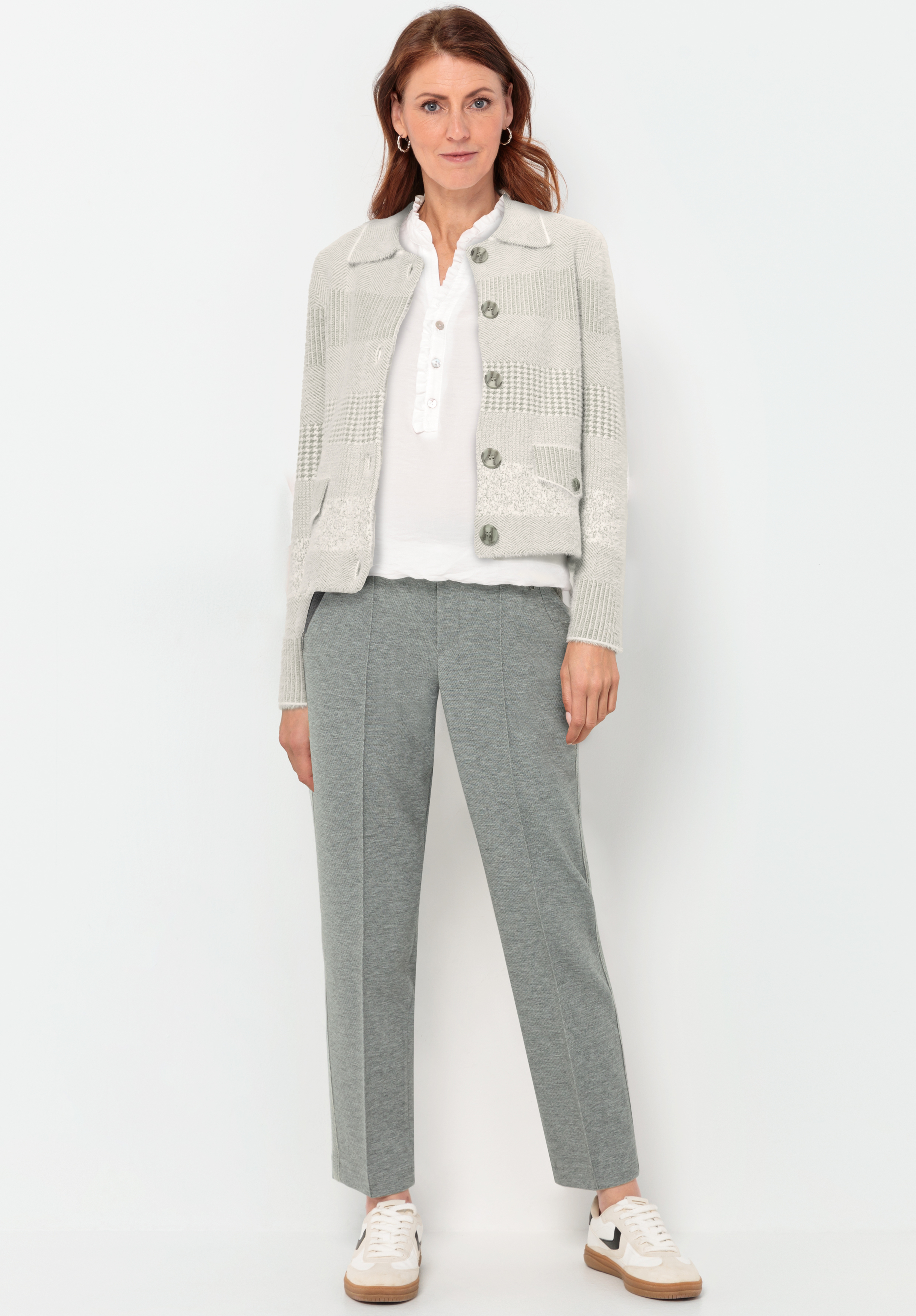 Eine Frau steht vor einem weißen Hintergrund und trägt eine hellkarierte Strickjacke, ein weißes Hemd mit Kragen, eine graue maßgeschneiderte Hose und weiße Turnschuhe. Sie hat langes braunes Haar und einen entspannten Gesichtsausdruck.