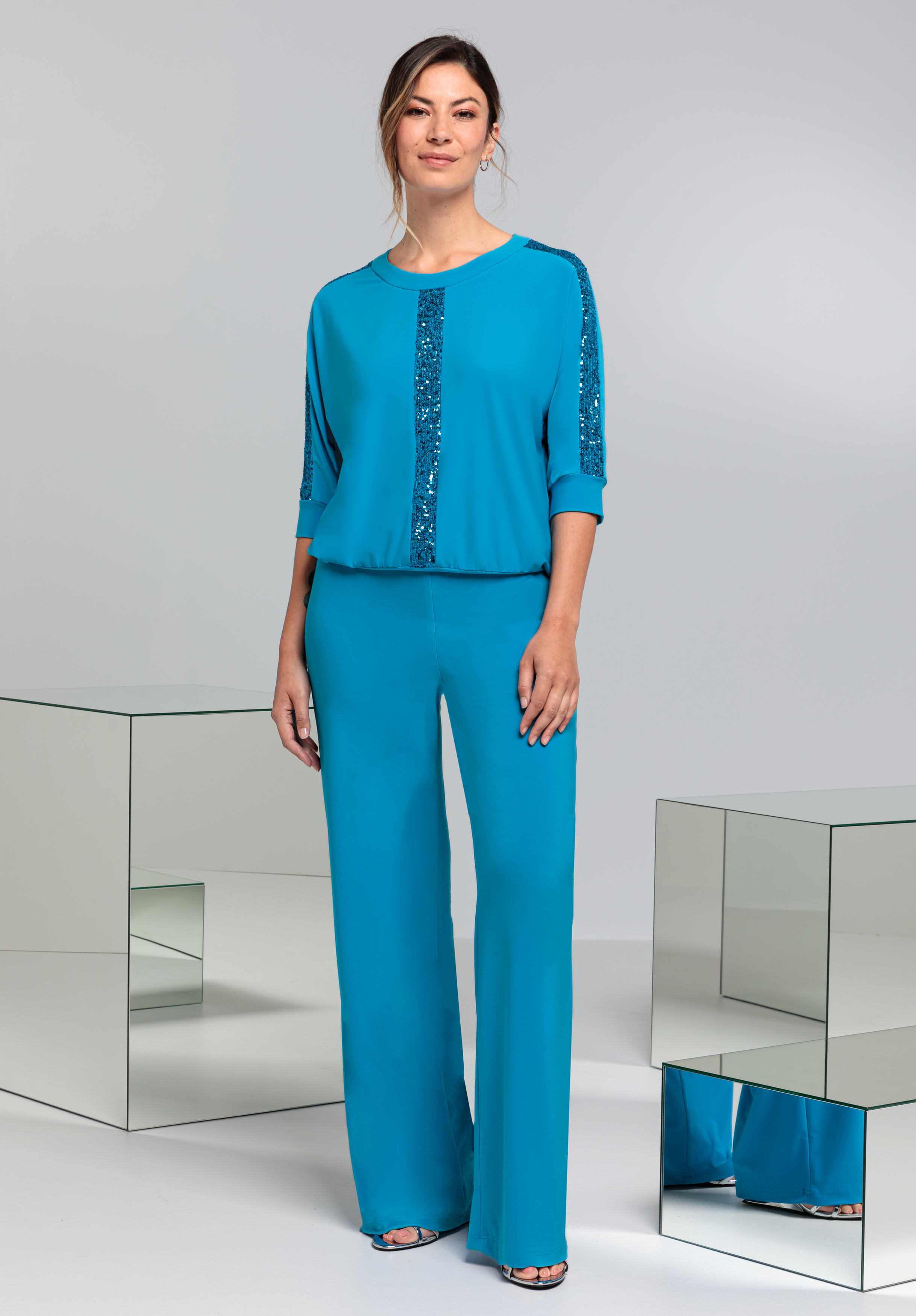 Eine lächelnde Frau in einem blauen Outfit mit Paillettendetails am Oberteil und an den Hosensäumen steht umgeben von verspiegelten Würfeln in einem minimalistischen Studio-Setting.