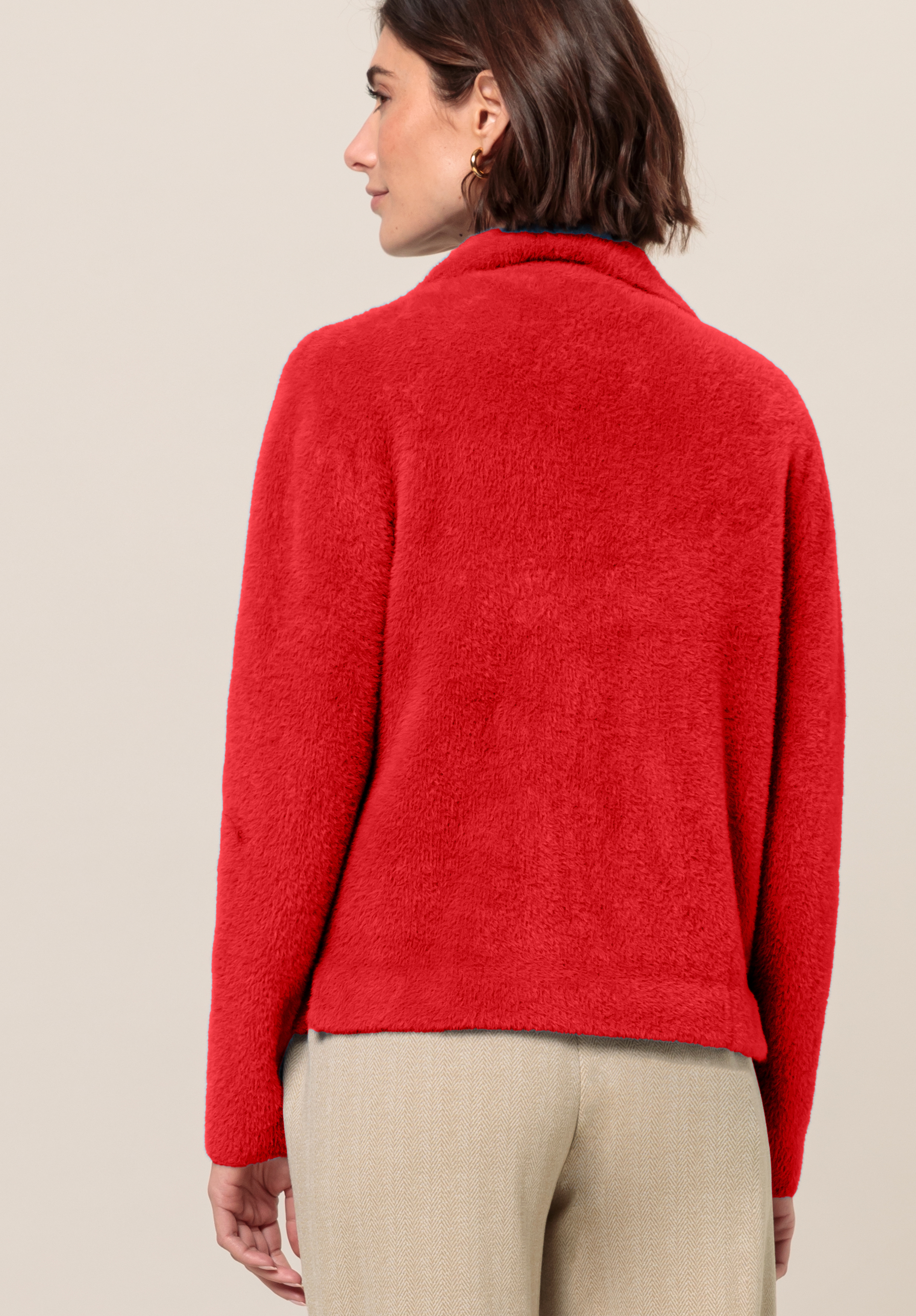 Von hinten ist eine Frau mit kurzen braunen Haaren zu sehen, die eine leuchtend rote, flauschige Jacke und beige Hosen trägt und vor einem schlichten hellbeigen Hintergrund steht.