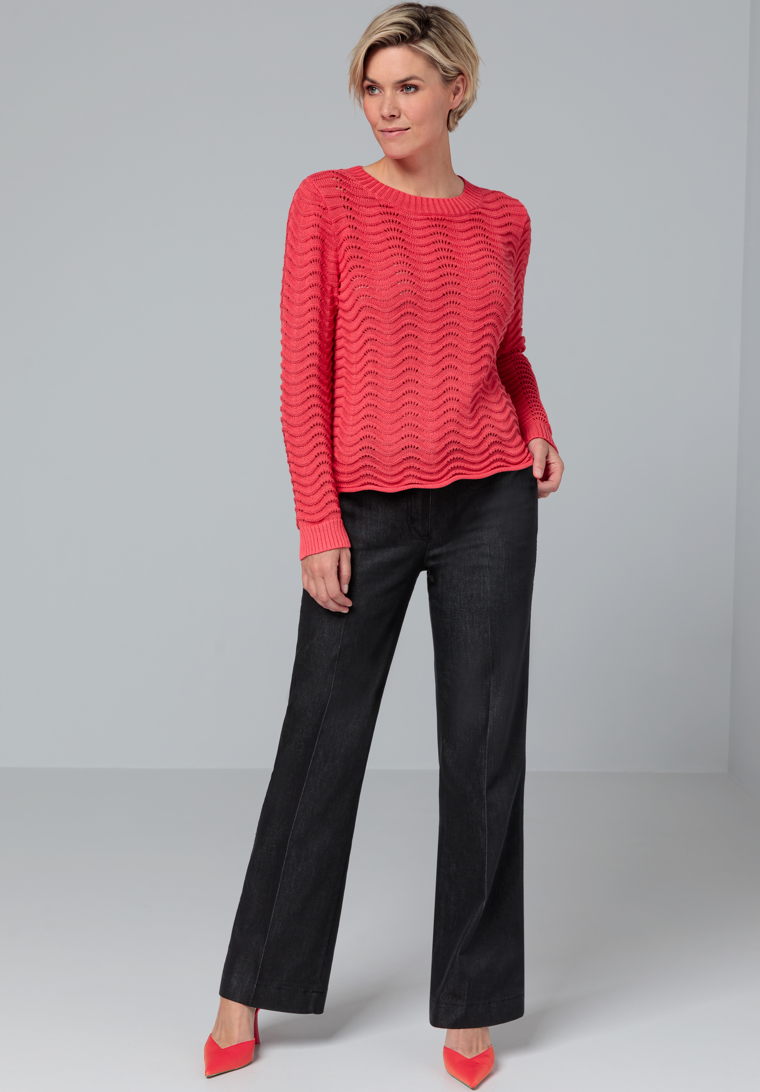 Eine Frau steht in einem Studio. Sie trägt einen strukturierten Korallenpullover, eine schwarze Hose mit weitem Bein und dazu passende Korallenschuhe. Sie hat eine Hand in der Tasche und lächelt leicht. Der Hintergrund ist schlicht hellgrau.