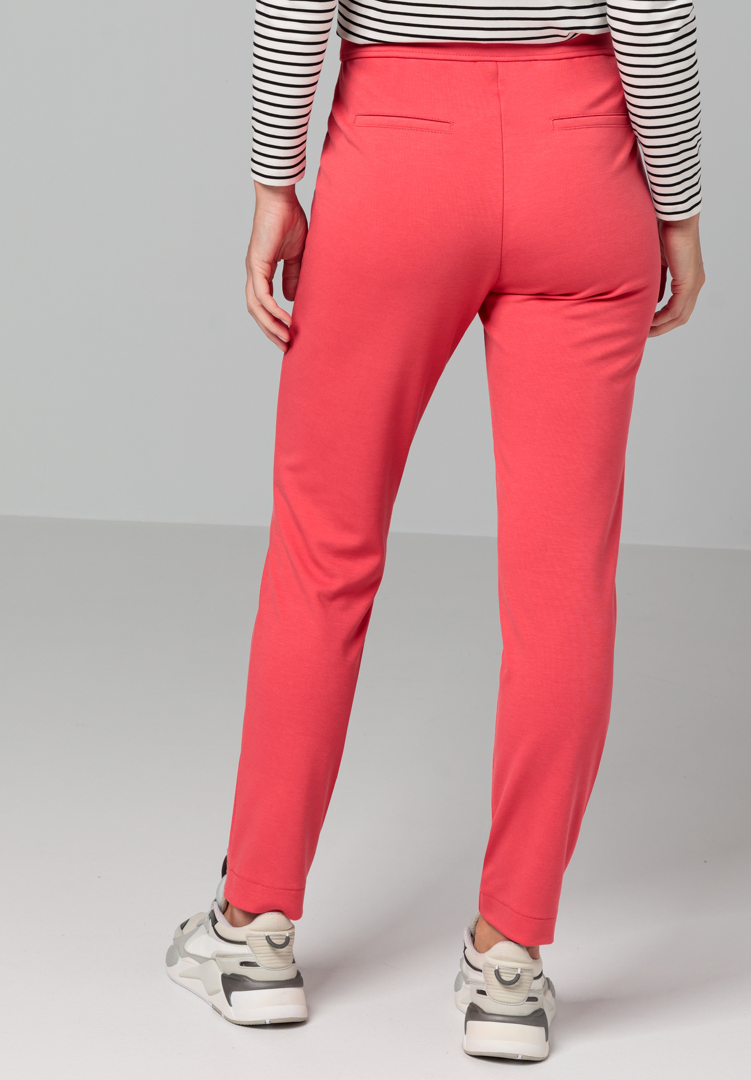 Eine Person in leuchtend rosa Hosen, einem schwarz-weiß gestreiften Langarmshirt und weißen Turnschuhen steht mit dem Rücken zur Kamera. Der Hintergrund ist schlicht und hellgrau.