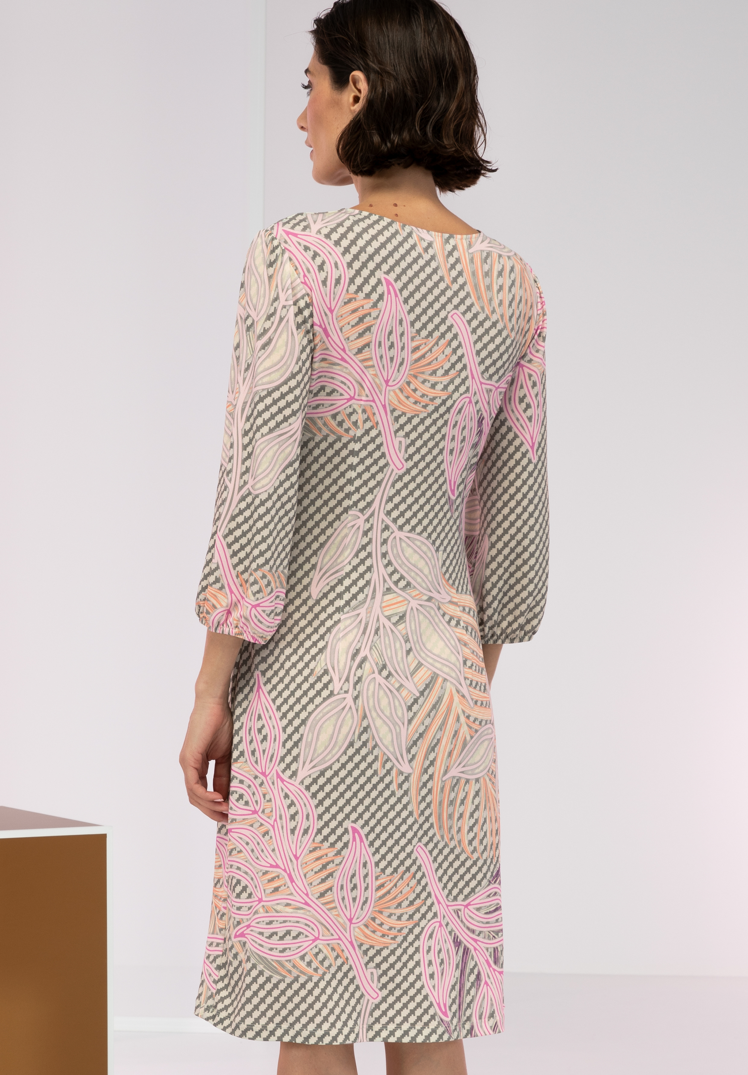 Eine Frau mit kurzen braunen Haaren trägt ein knielanges, gemustertes Kleid mit langen Ärmeln. Das Kleid hat einen geometrischen Hintergrund mit großen rosa, pfirsichfarbenen und weißen Blattmustern. Sie blickt von der Kamera ab.