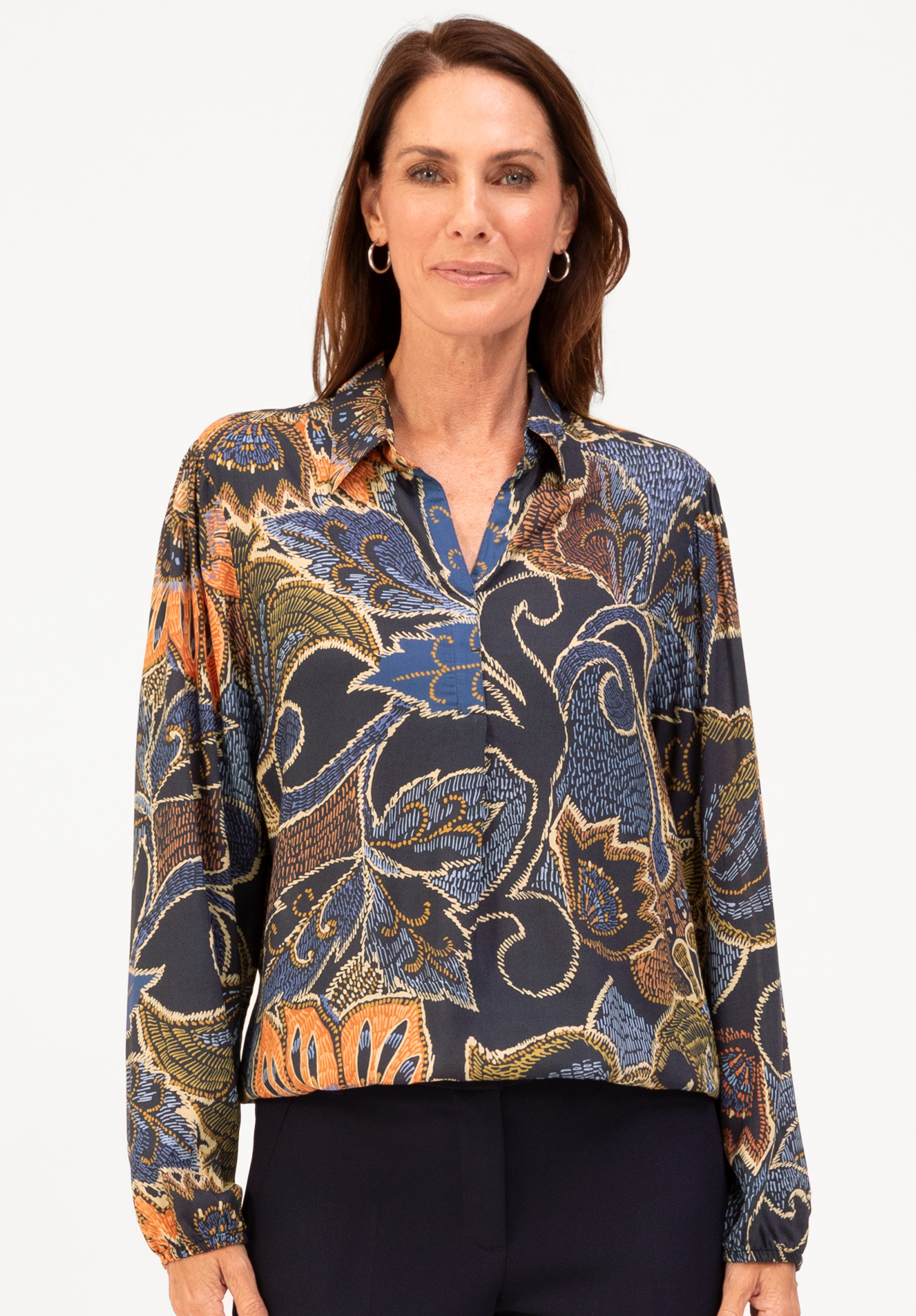 Eine Frau mit langen braunen Haaren, die goldene Ohrringe und eine langärmelige Kragenbluse mit einem bunten Blumen- und Blattmuster in Blau-, Orange- und Gelbtönen trägt, steht vor einem schlichten weißen Hintergrund.