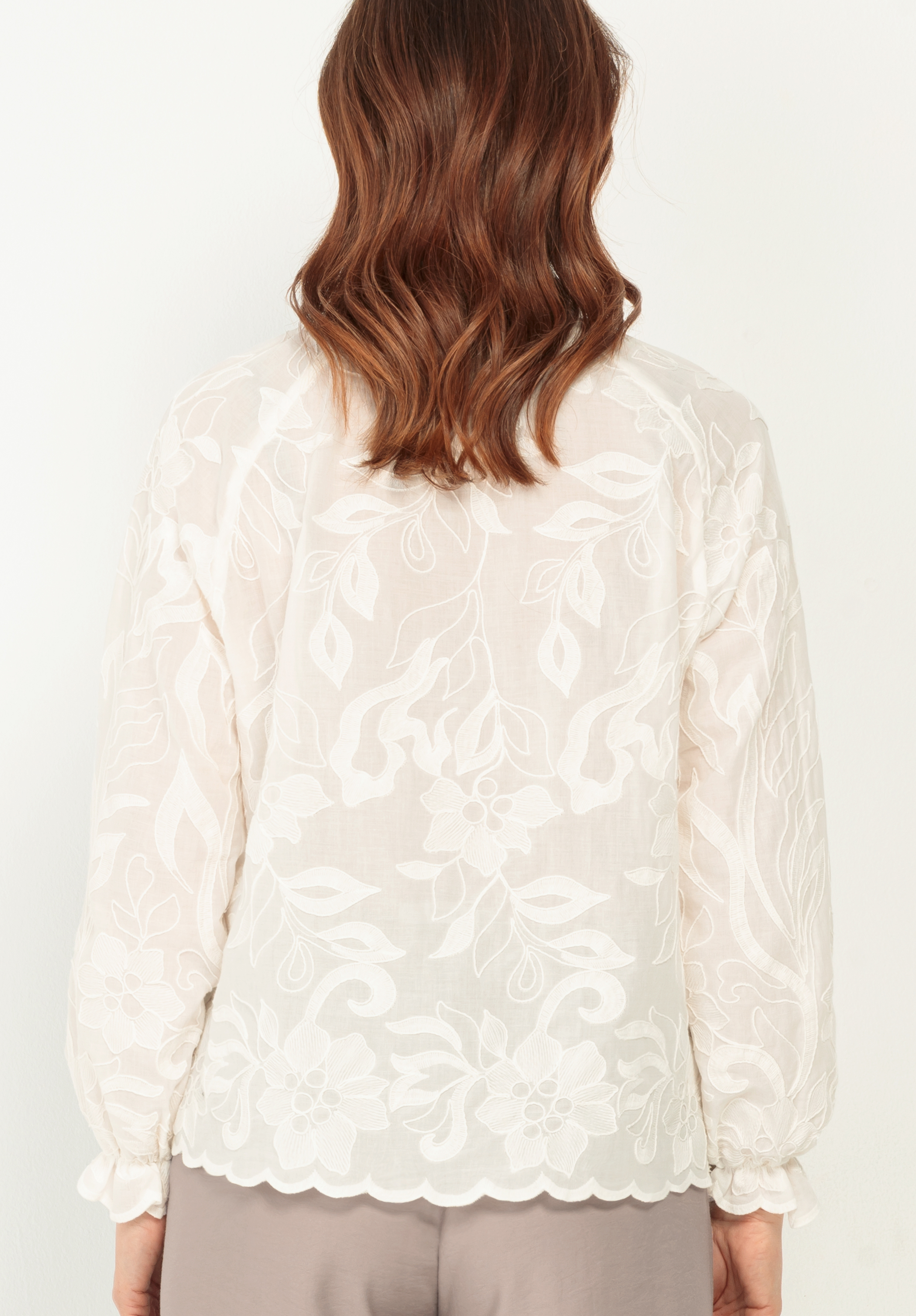 Eine Person mit gewelltem, schulterlangem braunem Haar ist von hinten zu sehen. Sie trägt eine durchsichtige, langärmelige weiße Bluse mit Blumenstickerei und geschwungenem Saum, dazu eine helle Hose.