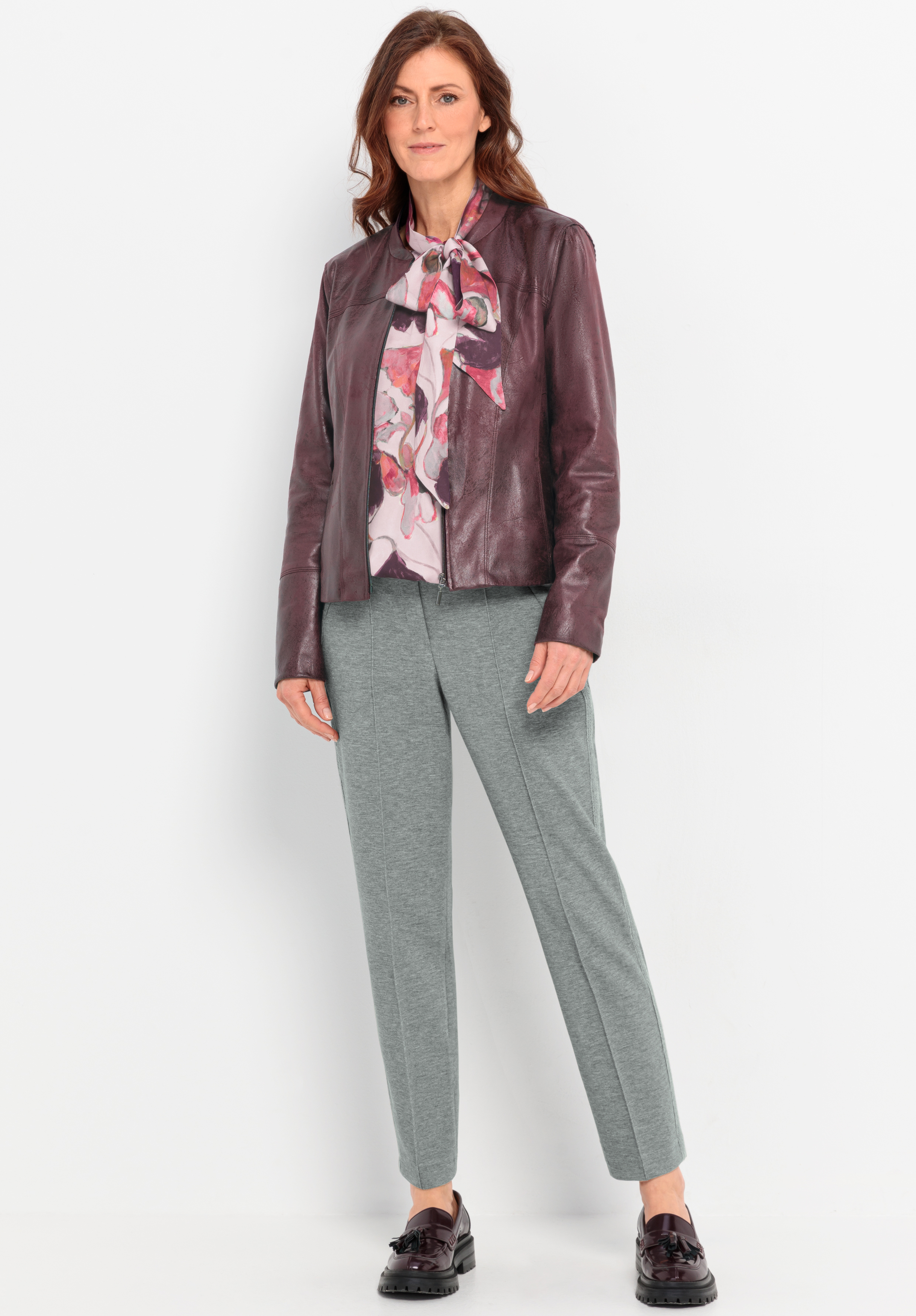 Eine Frau steht vor einem weißen Hintergrund und trägt eine weinrote Lederjacke, eine geblümte Bluse mit einer Schleife am Kragen, eine graue Hose und dunkle Halbschuhe. Sie hat gewelltes braunes Haar und ein leichtes Lächeln.