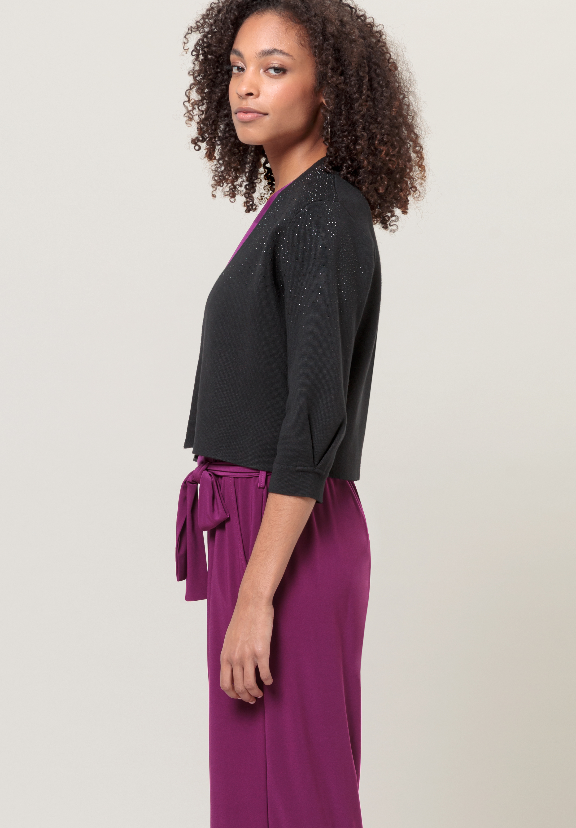 Eine Frau mit lockigem Haar steht seitlich vor einem schlichten hellen Hintergrund. Sie trägt eine schwarze Strickjacke mit Dreiviertelärmeln und dezentem Glitzer und ein violettes Kleid mit Taillenband.