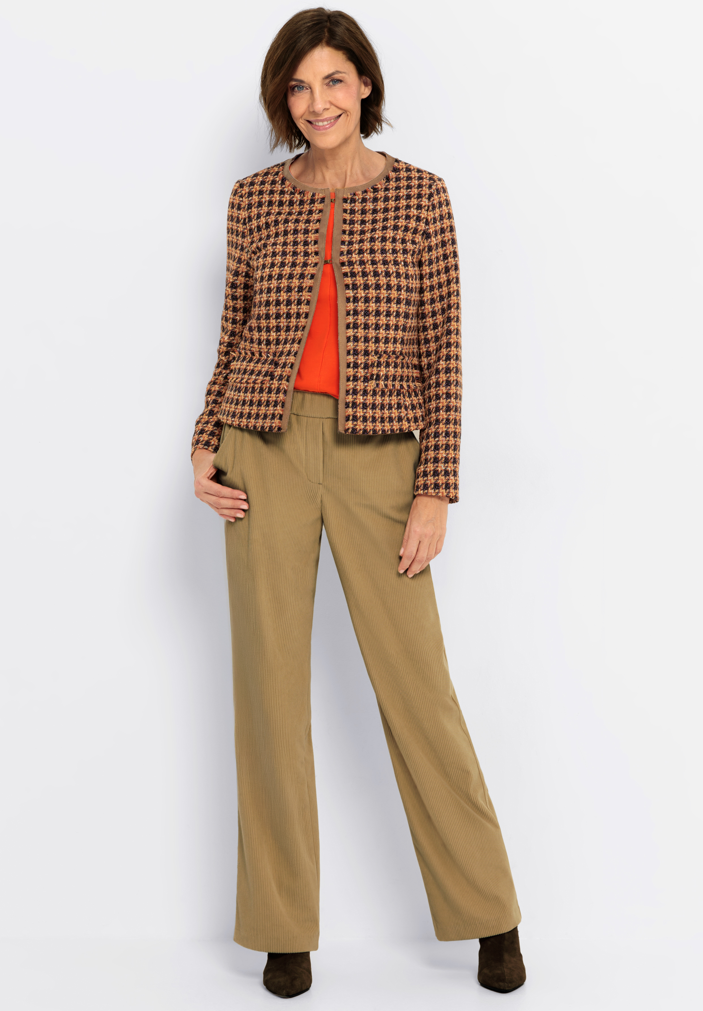 Eine Frau steht lächelnd in einer braun-schwarzen Jacke mit Hahnentrittmuster über einem orangefarbenen Oberteil, einer hellbraunen Hose mit weitem Bein und dunklen Schuhen vor einem schlichten weißen Hintergrund.