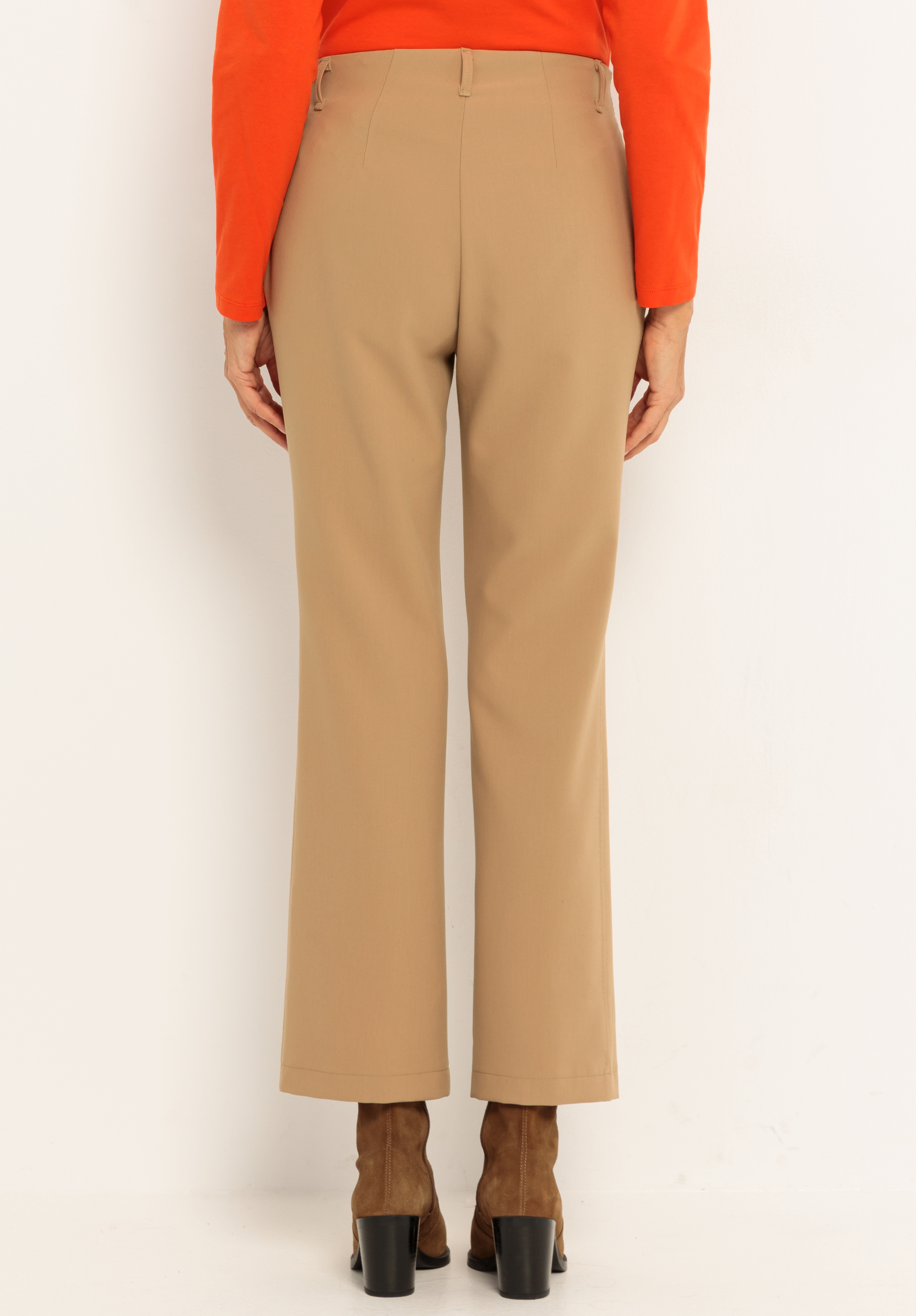 Eine von der Kamera abgewandte Person trägt eine hellbraune Hose mit geradem Bein, ein orangefarbenes, langärmeliges Oberteil und braune, hochhackige Stiefel und steht vor einem grauweißen Hintergrund.