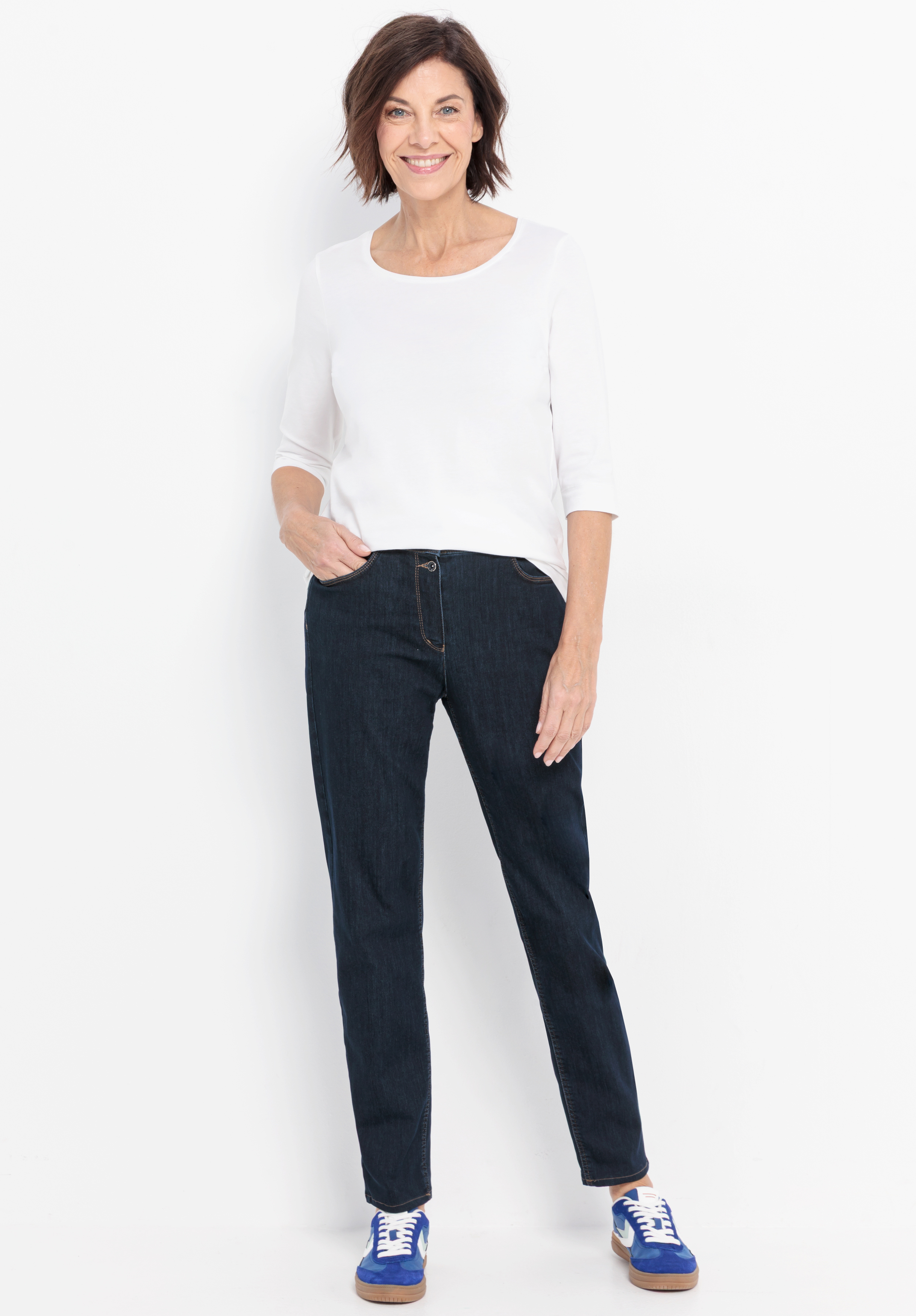 Eine Frau mit kurzen braunen Haaren lächelt, trägt ein weißes dreiviertelärmliges Oberteil, dunkle Jeans und blaue Turnschuhe und steht vor einem schlichten weißen Hintergrund.