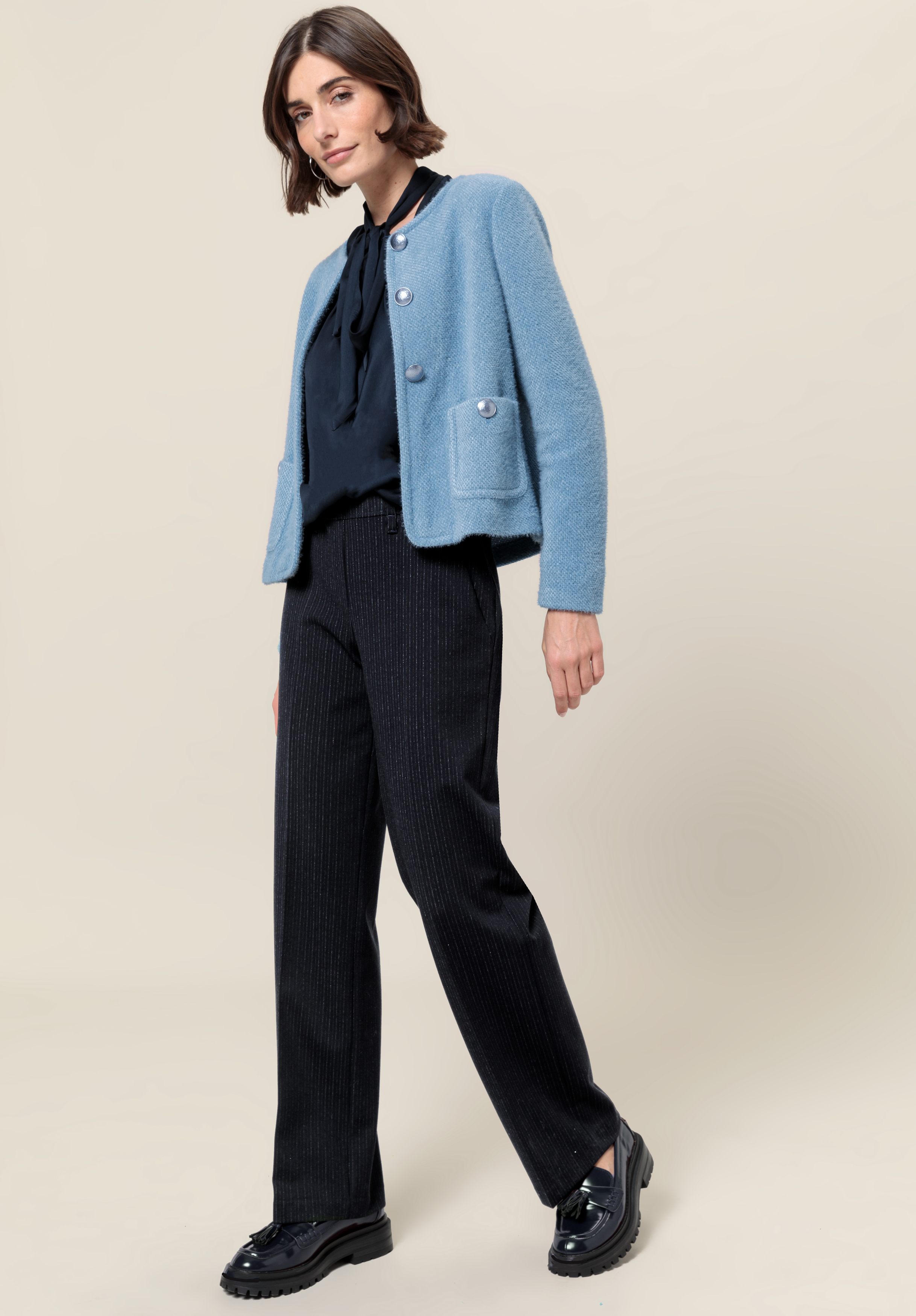 Eine Frau steht vor einem hellen Hintergrund. Sie trägt eine hellblaue Strickjacke mit Knöpfen, eine schwarze Bluse mit Fliege, eine schwarze Nadelstreifenhose und schwarze Slipper. Sie hat kurze braune Haare und eine entspannte, selbstbewusste Pose.