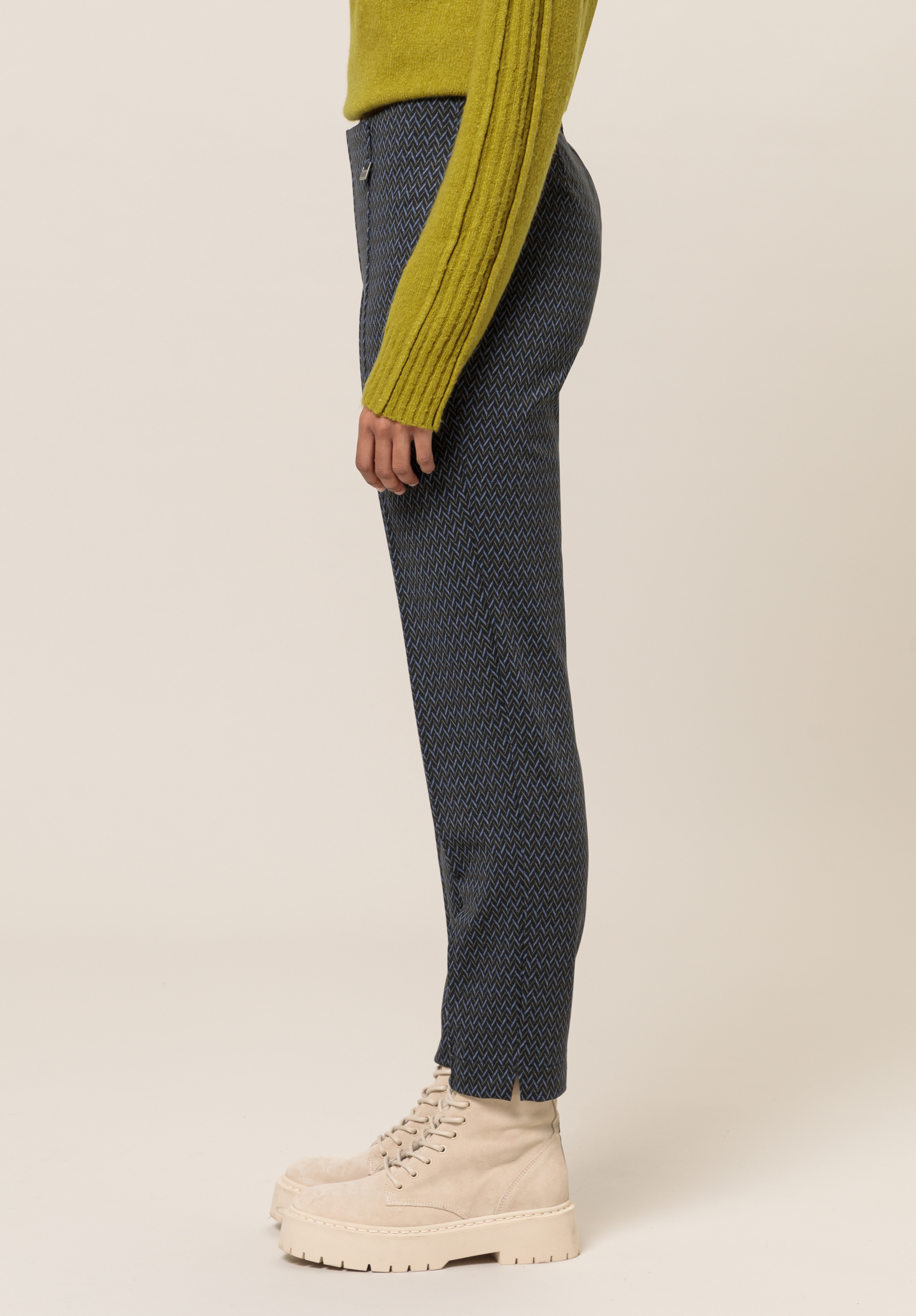 Eine Person steht im Profil und trägt einen strukturierten grünen Pullover, blau gemusterte Hosen und beige Plateaustiefel mit Schnürung vor einem schlichten hellen Hintergrund.