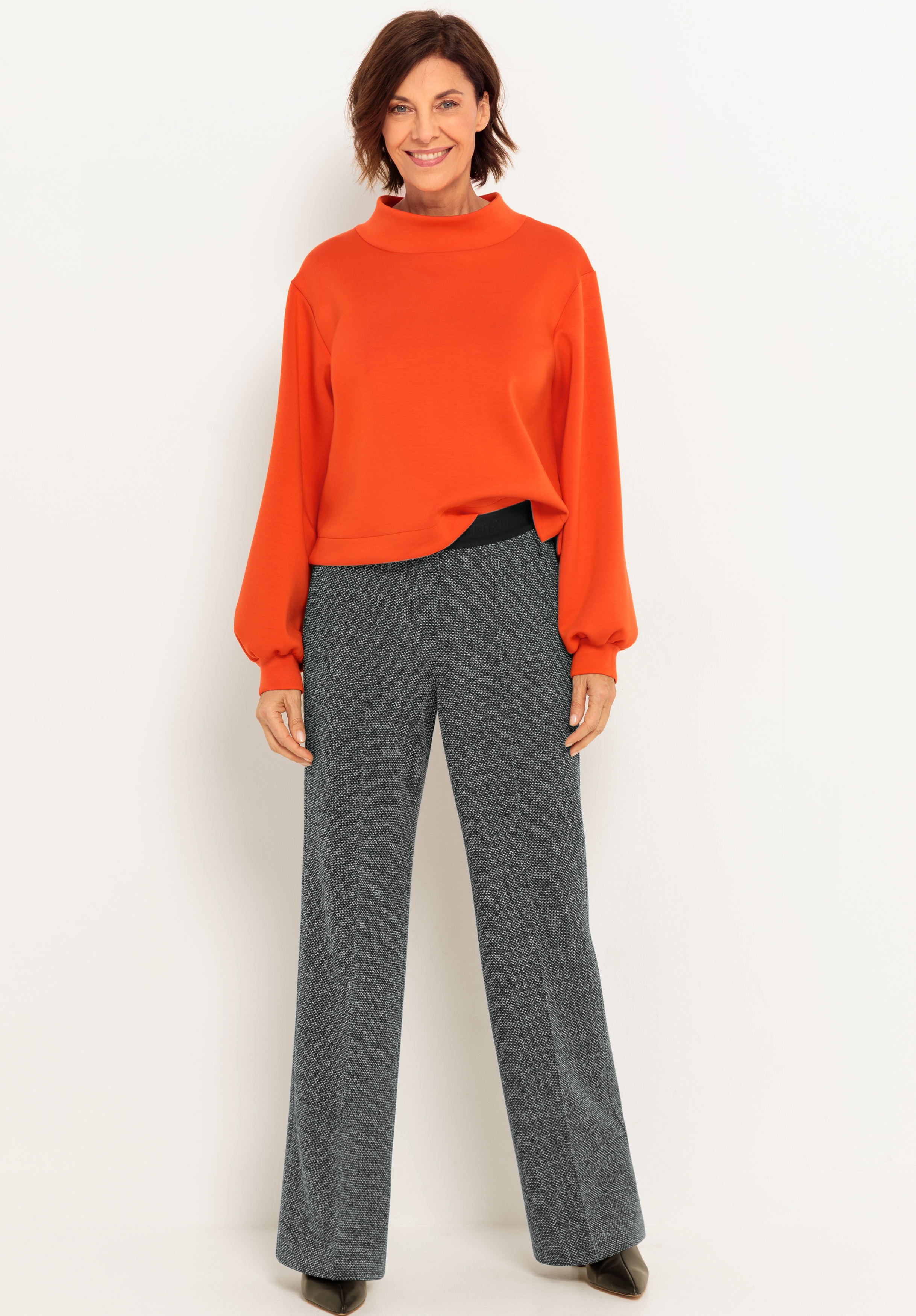 Eine Frau steht lächelnd vor einem weißen Hintergrund und trägt ein orangefarbenes langärmeliges Oberteil, eine graue Hose mit weitem Bein und braune Schuhe.