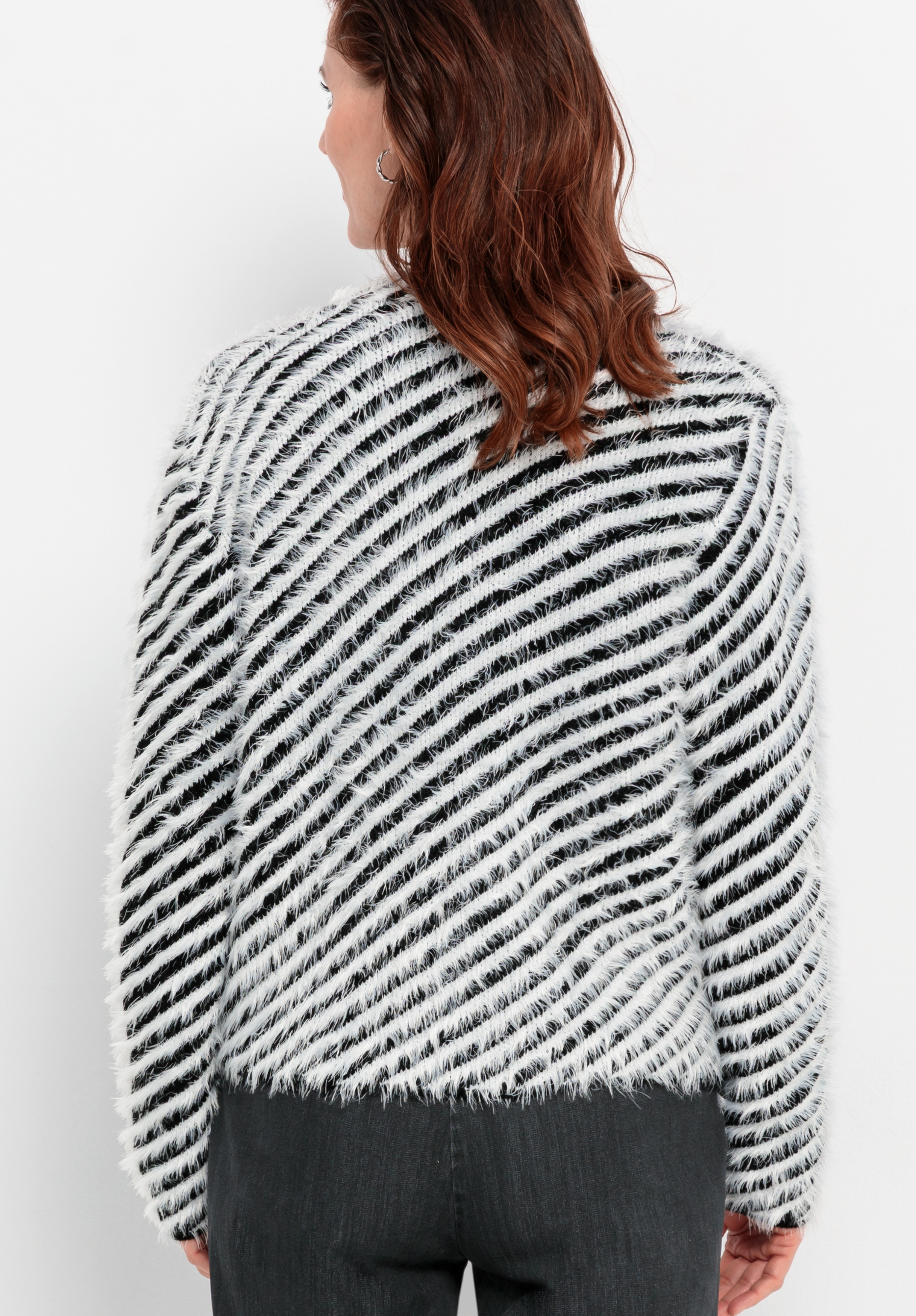 Eine Person mit welligem braunem Haar ist von hinten zu sehen; sie trägt einen flauschigen schwarz-weiß gestreiften Pullover und eine dunkle Hose und steht vor einem schlichten weißen Hintergrund.