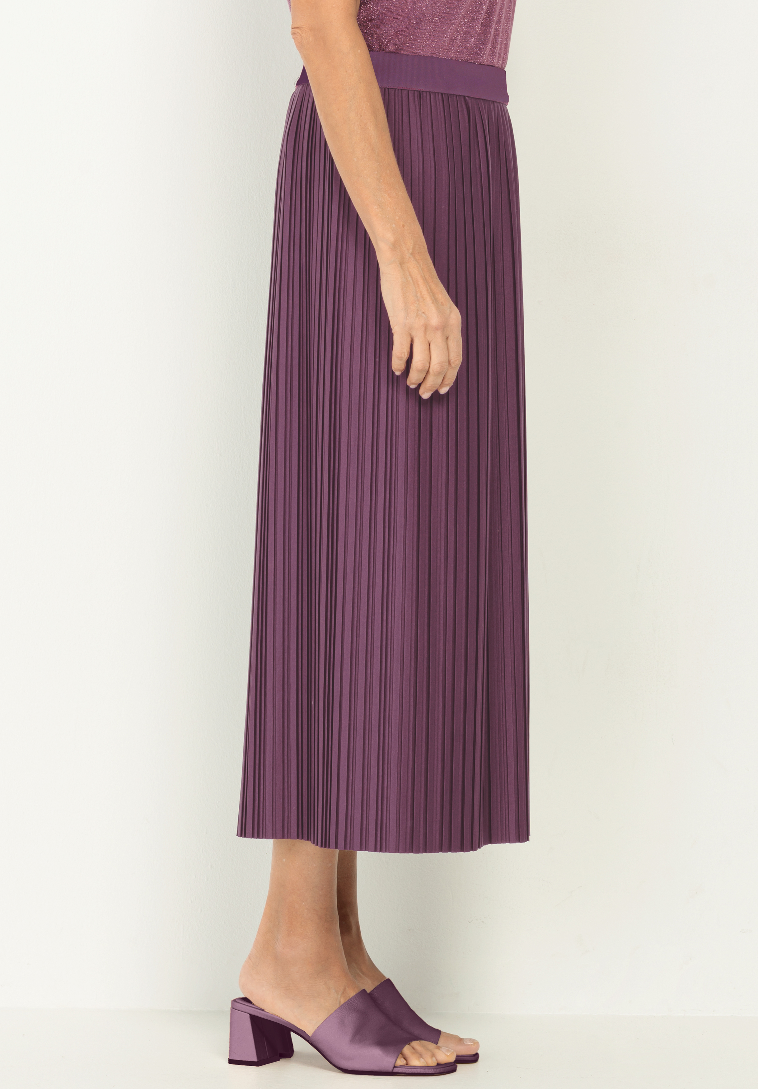 Eine Person, die einen lilafarbenen plissierten Midirock und passende lilafarbene hochhackige Sandalen trägt, steht vor einem schlichten weißen Hintergrund, von der Seite gesehen.