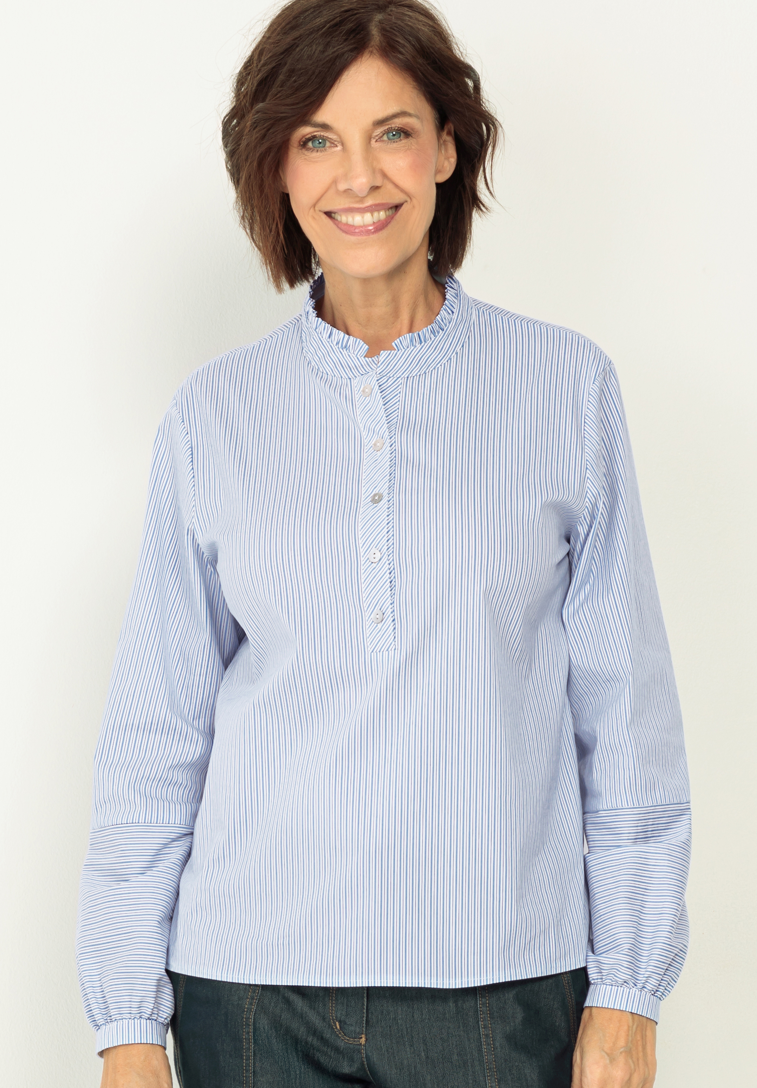 Schlupfbluse ADINA Eine Frau mit kurzen braunen Haaren lächelt, während sie eine blau-weiß gestreifte Bluse mit Rüschenkragen und langen Ärmeln vor einem schlichten hellen Hintergrund trägt.