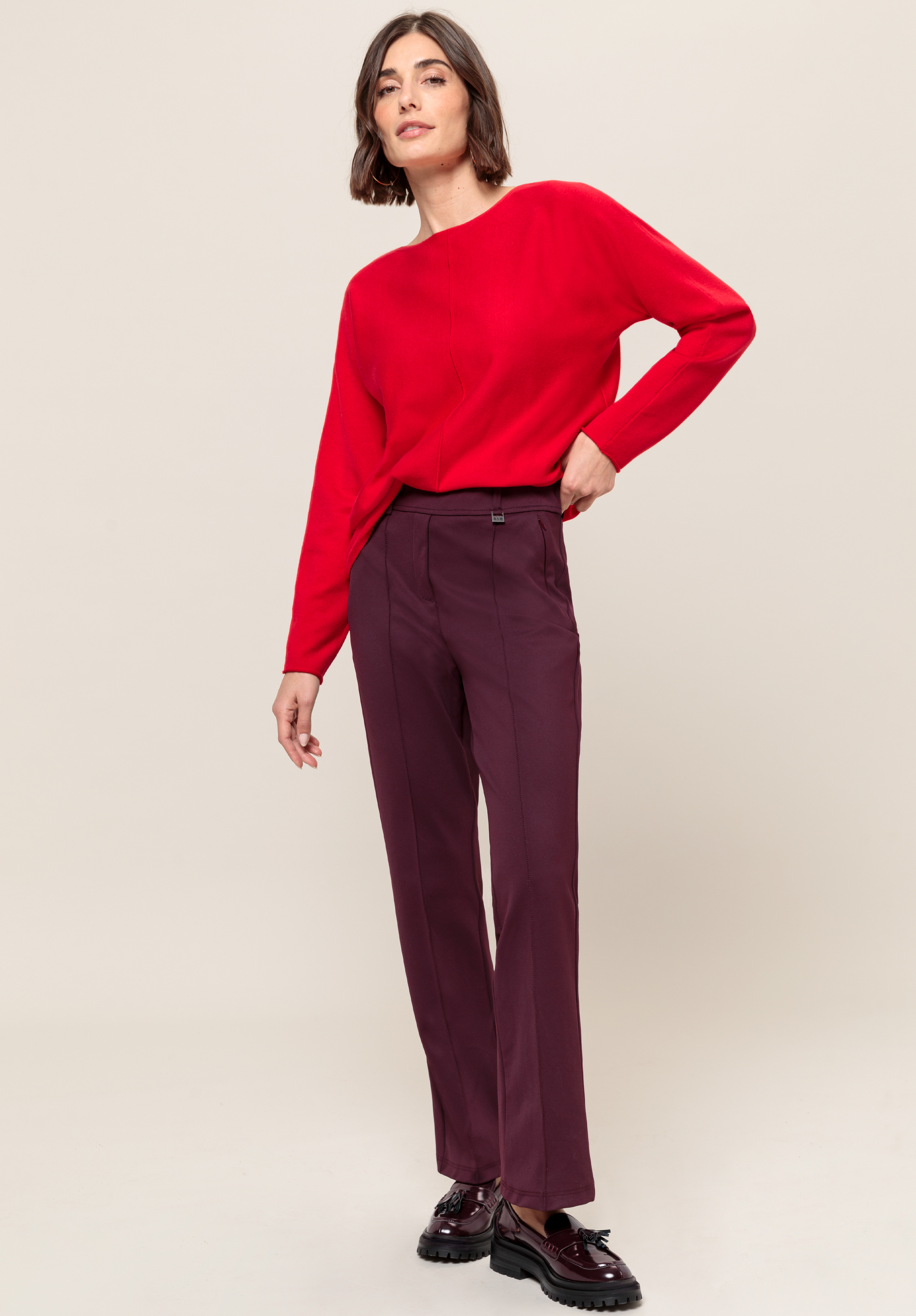 Eine Frau steht vor einem schlichten Hintergrund. Sie trägt ein leuchtend rotes Langarmshirt, eine burgunderfarbene Hose und dunkelburgunderfarbene Plateauschuhe.