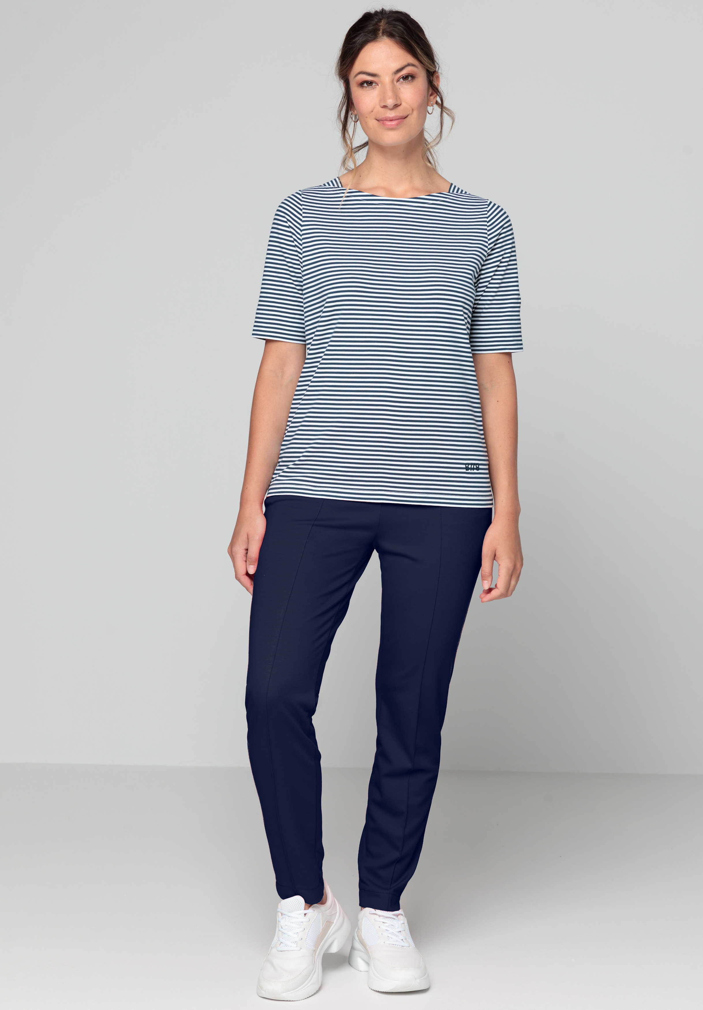 Eine Frau steht vor einem schlichten Hintergrund. Sie trägt ein marineblau-weiß gestreiftes Kurzarmoberteil, eine dunkelblaue Hose und weiße Turnschuhe. Sie lächelt sanft und lässt ihre Hände entspannt an den Seiten hängen.