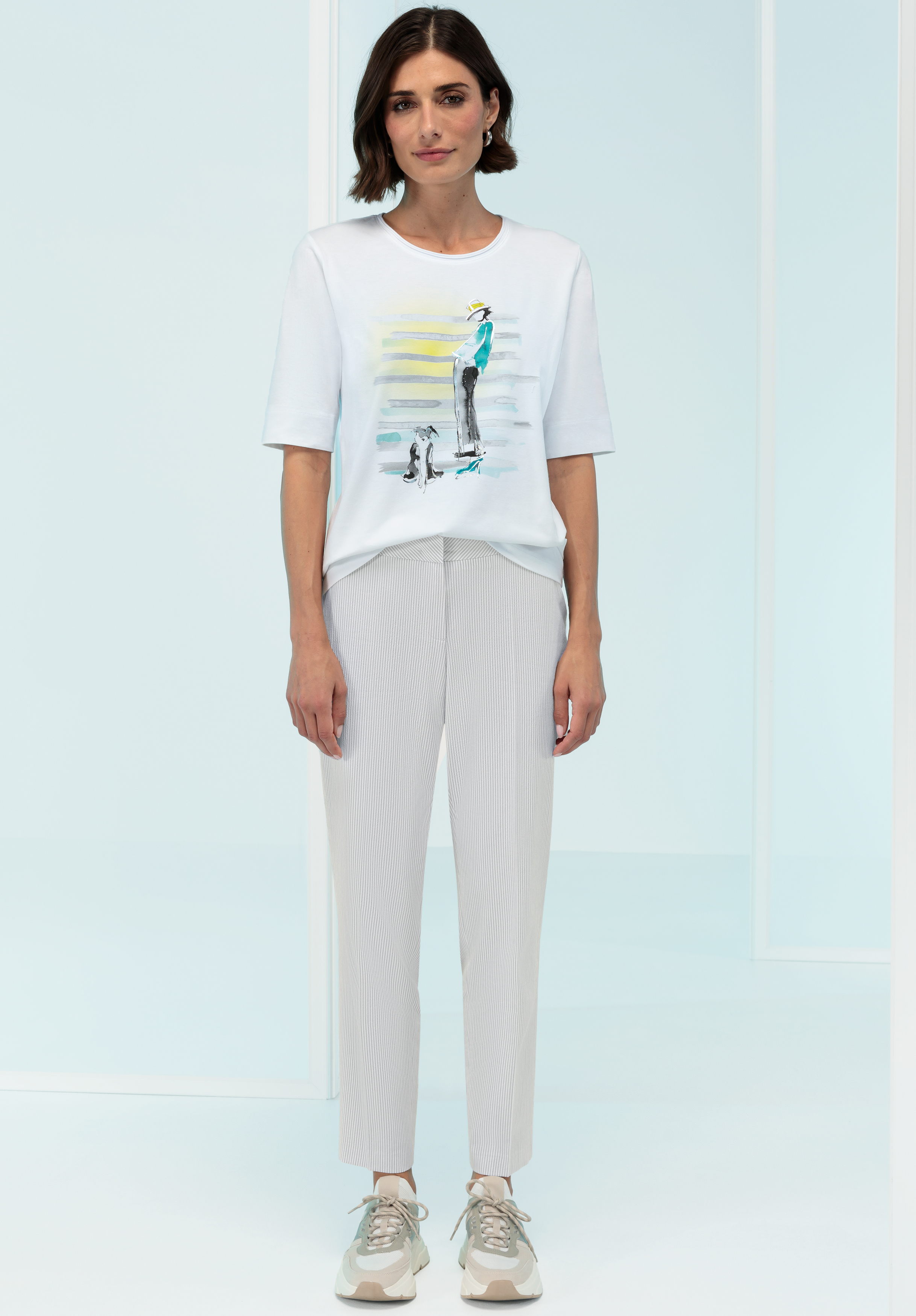 Eine Frau mit kurzen braunen Haaren trägt ein weißes T-Shirt mit Aufdruck, hellgraue knöchellange Hosen und beige Turnschuhe und steht in einem minimalistischen, hellen Studio.