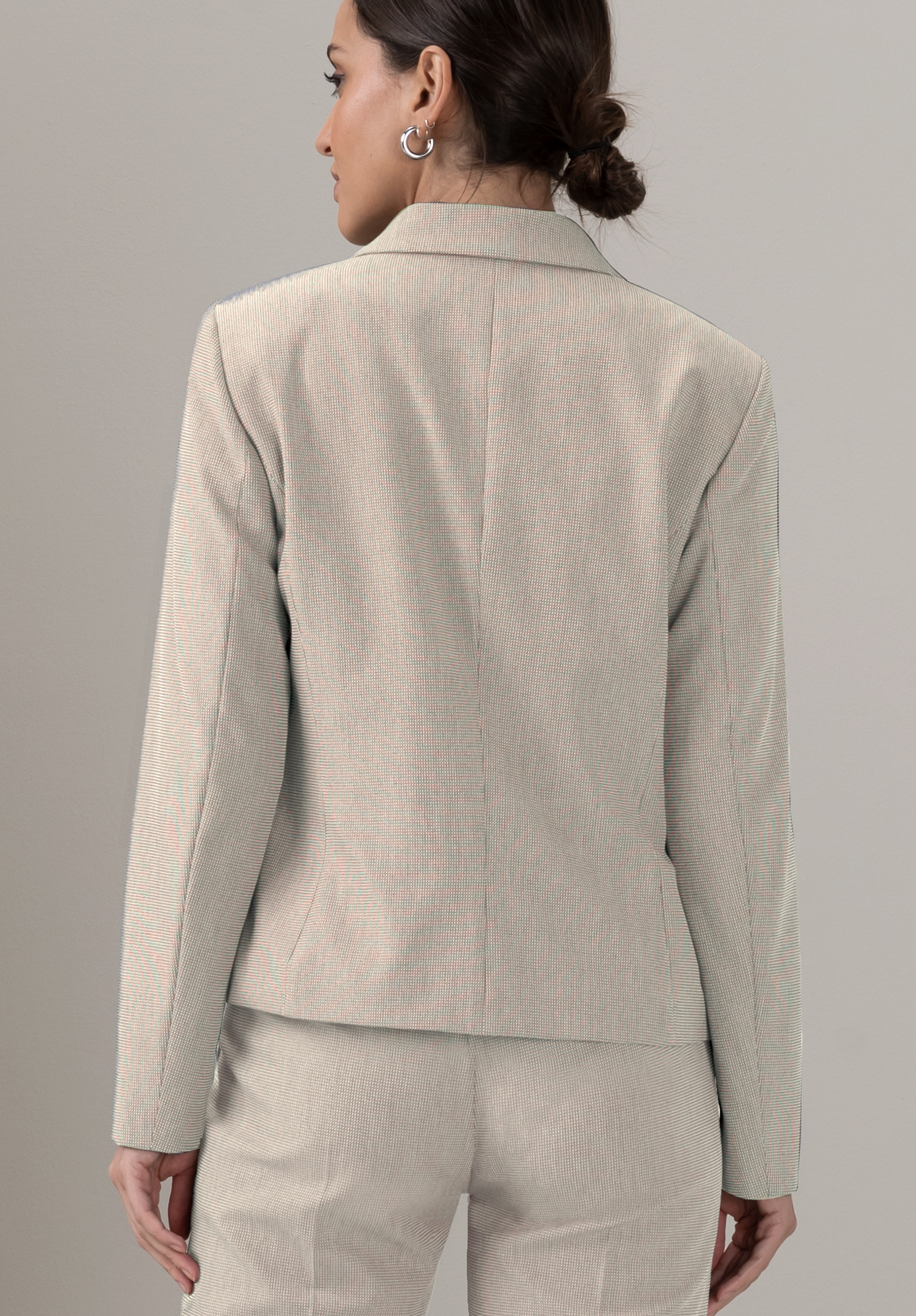 Eine Frau mit zurückgebundenem braunem Haar, die einen hellbeigen Blazer und eine passende Hose trägt, steht mit dem Rücken zur Kamera vor einem schlichten, neutralen Hintergrund.