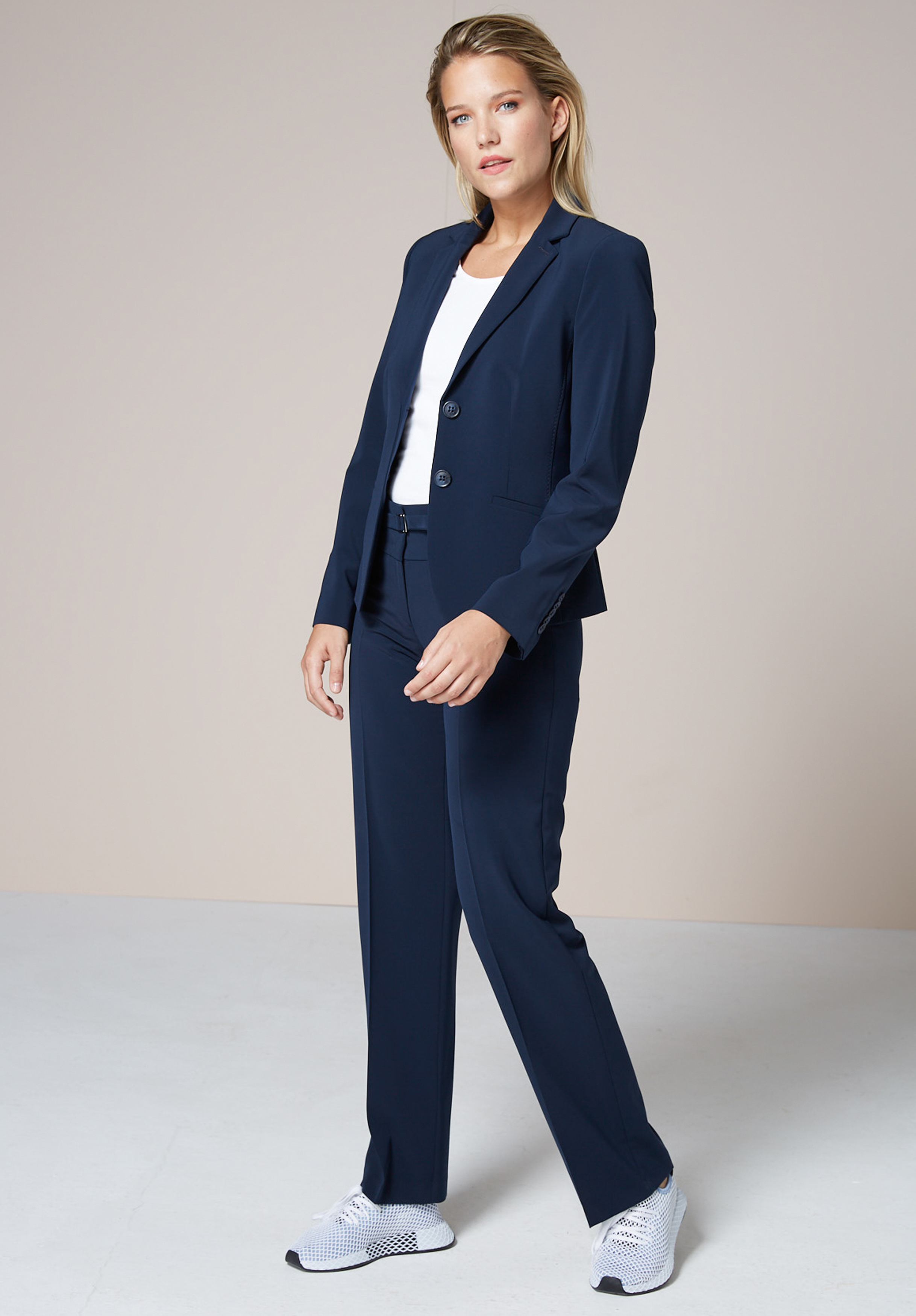 Eine Frau steht in einem Studio und trägt einen marineblauen Hosenanzug, ein weißes Top und weiße Turnschuhe. Sie hat ihr blondes Haar zurückgesteckt und blickt leicht zur Seite, mit einem neutralen Hintergrund hinter ihr.