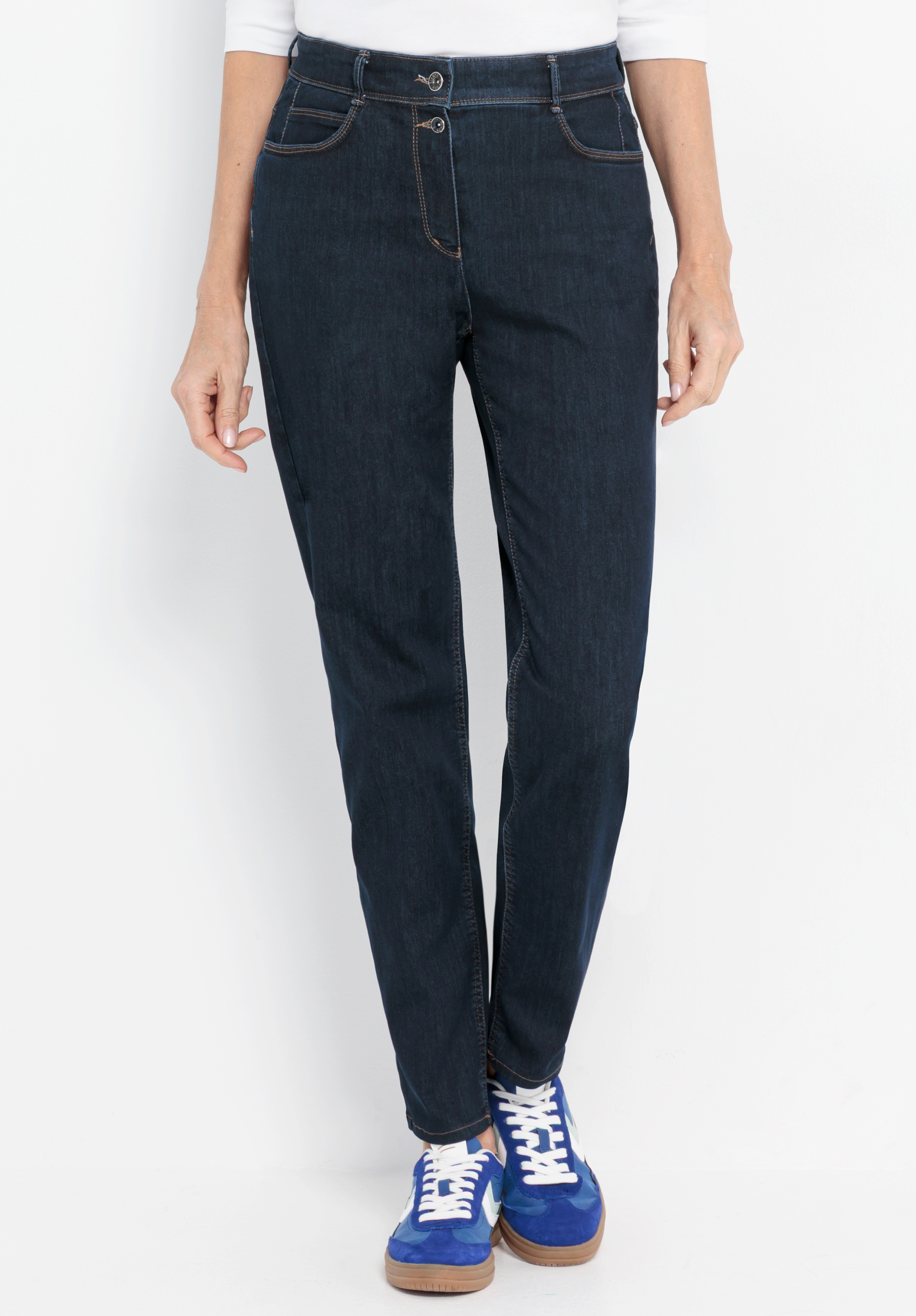 Eine Person, die dunkelblaue Jeans mit braunen Nähten, ein weißes Oberteil und blaue Turnschuhe trägt, steht vor einem schlichten weißen Hintergrund. Nur die untere Körperhälfte ist zu sehen.