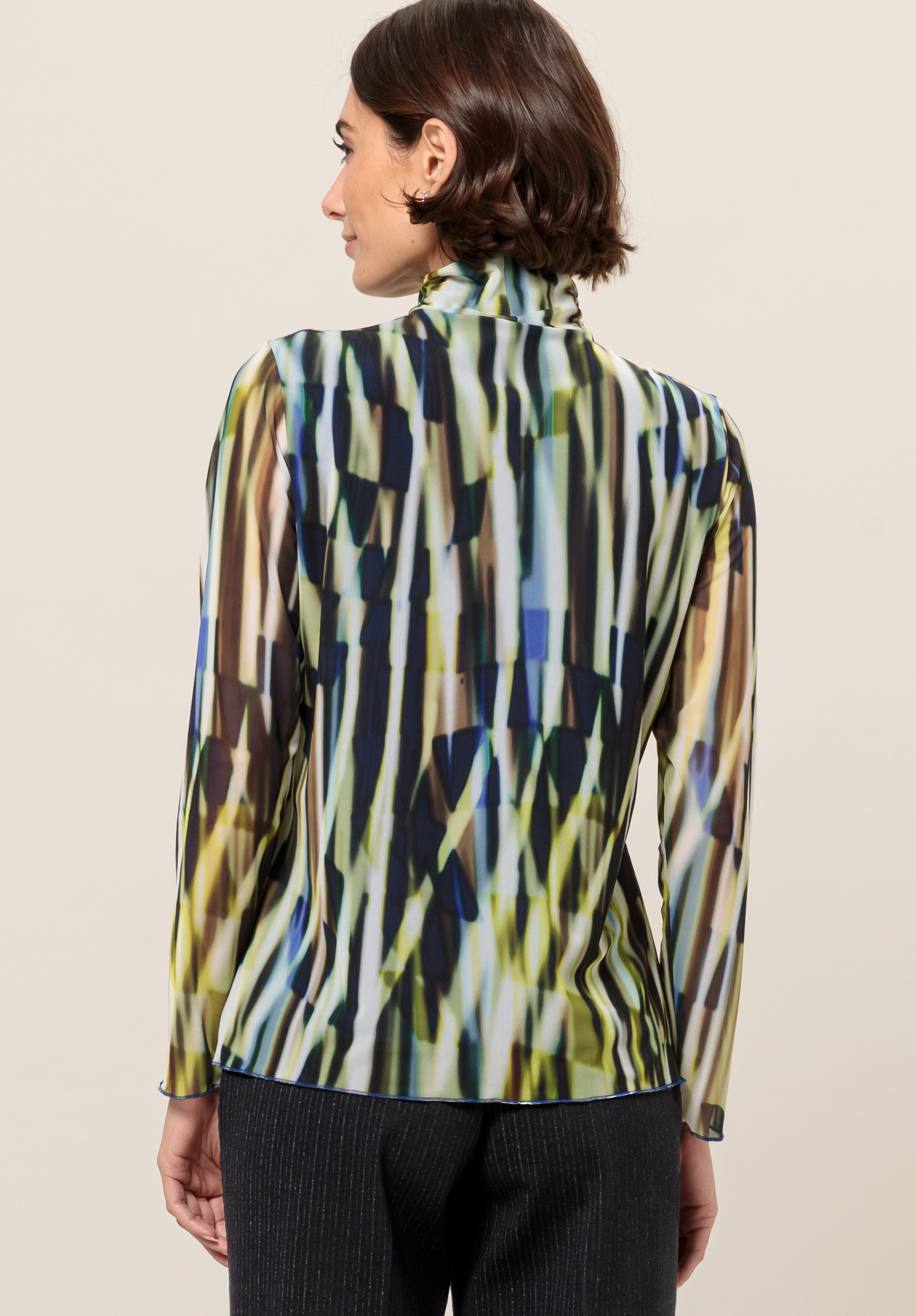 Von hinten ist eine Frau mit kurzen braunen Haaren zu sehen, die eine langärmlige Bluse mit hohem Kragen und abstraktem, mehrfarbigem Muster in Grün-, Blau-, Schwarz- und Gelbtönen sowie eine dunkle Hose trägt.