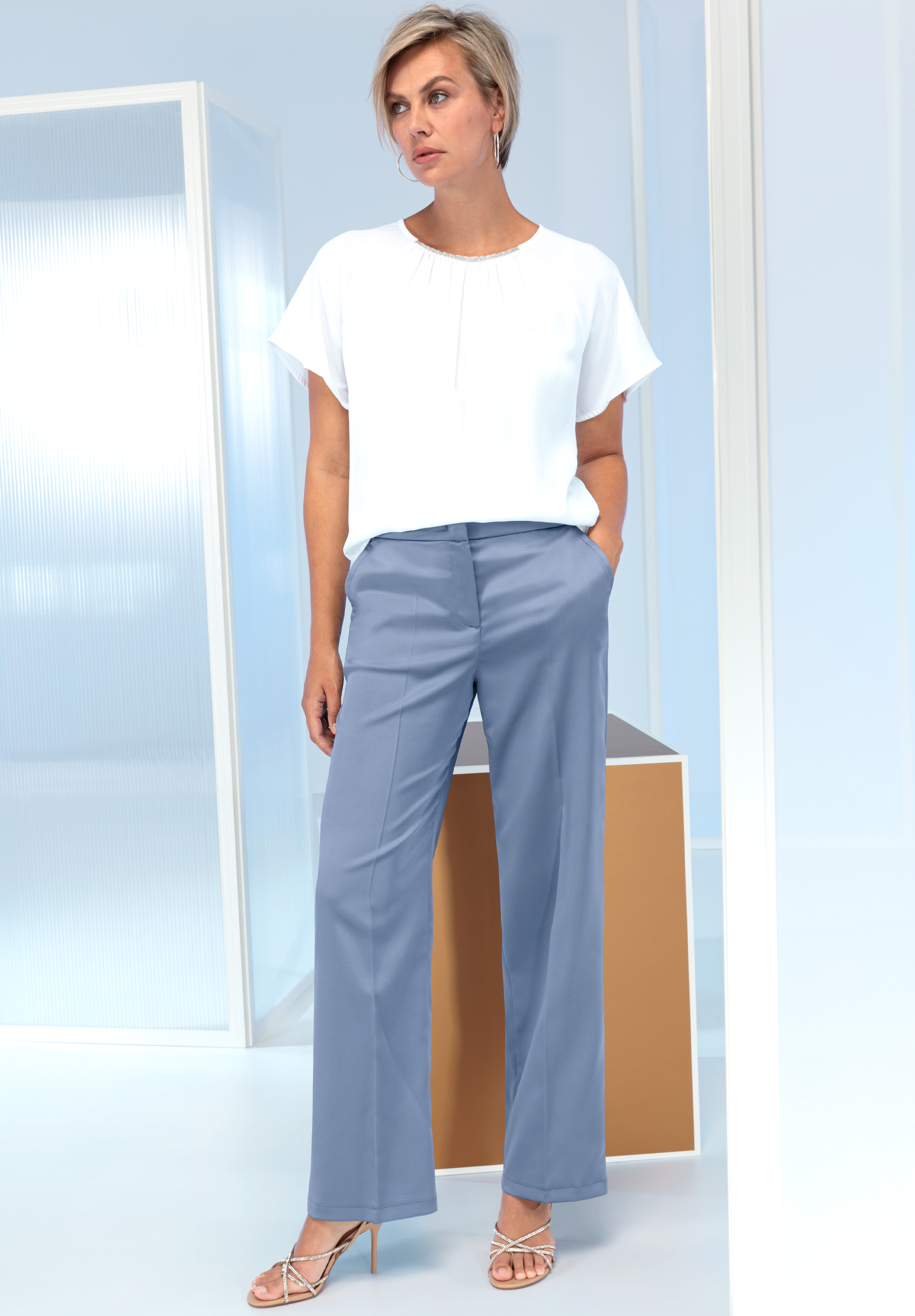 Eine Frau mit kurzen blonden Haaren steht drinnen. Sie trägt eine weite weiße Bluse, eine hellblaue Hose mit hoher Taille und Riemchensandalen. Sie hat die Hände in den Taschen und posiert vor einem minimalistischen, modernen Hintergrund.