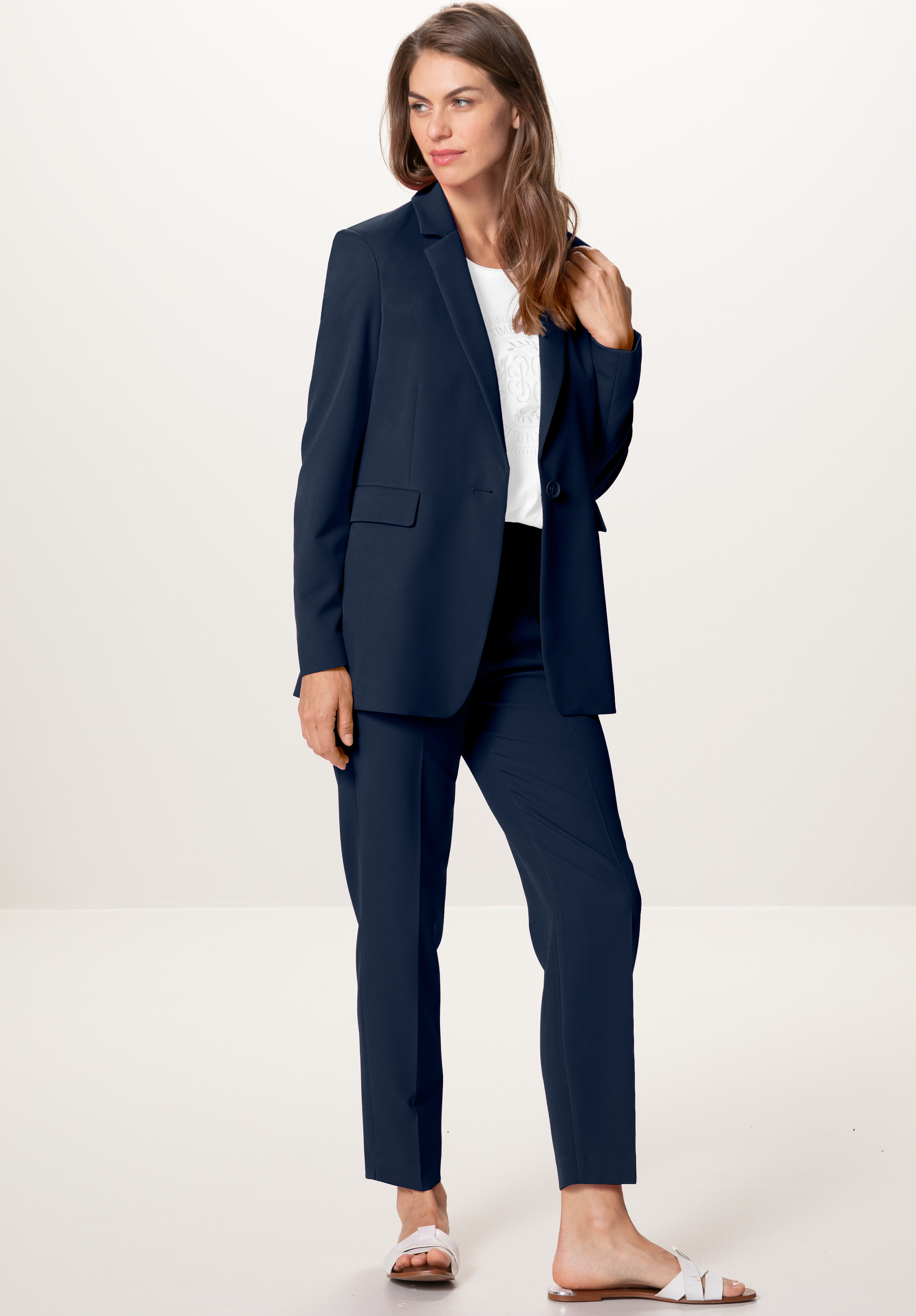 Eine Frau steht in einem marineblauen Hosenanzug mit einem weißen Oberteil und weißen Slide-Sandalen vor einem schlichten, hellen Hintergrund. Sie hat langes braunes Haar und blickt leicht zur Seite, wobei sie ihr Blazer-Revers festhält.