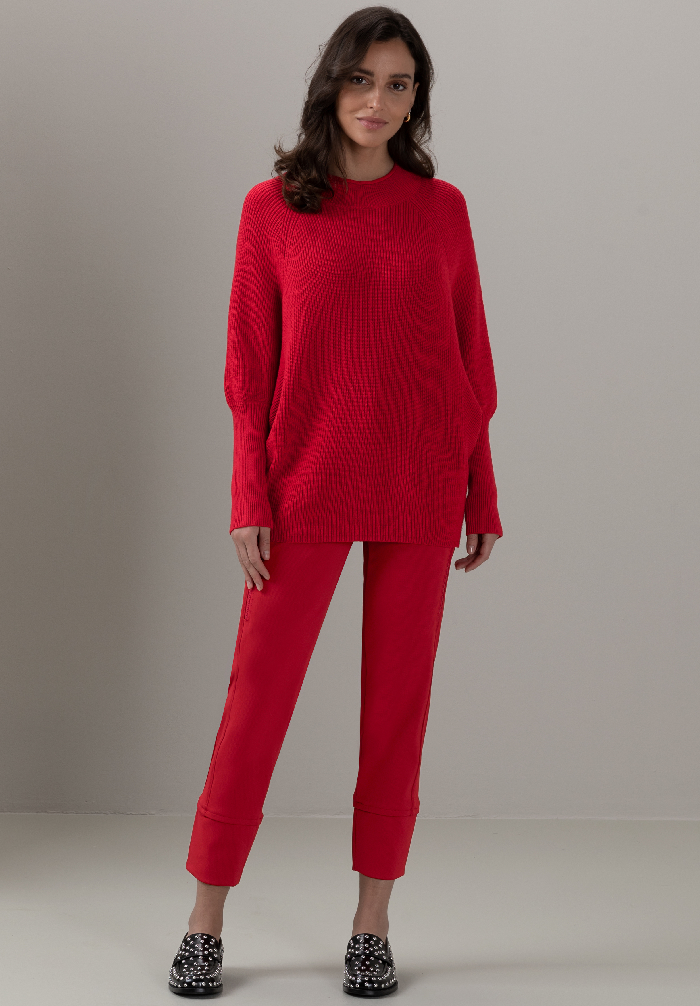 Eine Frau steht vor einem schlichten hellgrauen Hintergrund. Sie trägt einen locker sitzenden roten Pullover, eine dazu passende rote Hose mit Bündchen und schwarze Slipper mit weißen Punkten. Sie hat langes dunkles Haar und lächelt sanft.
