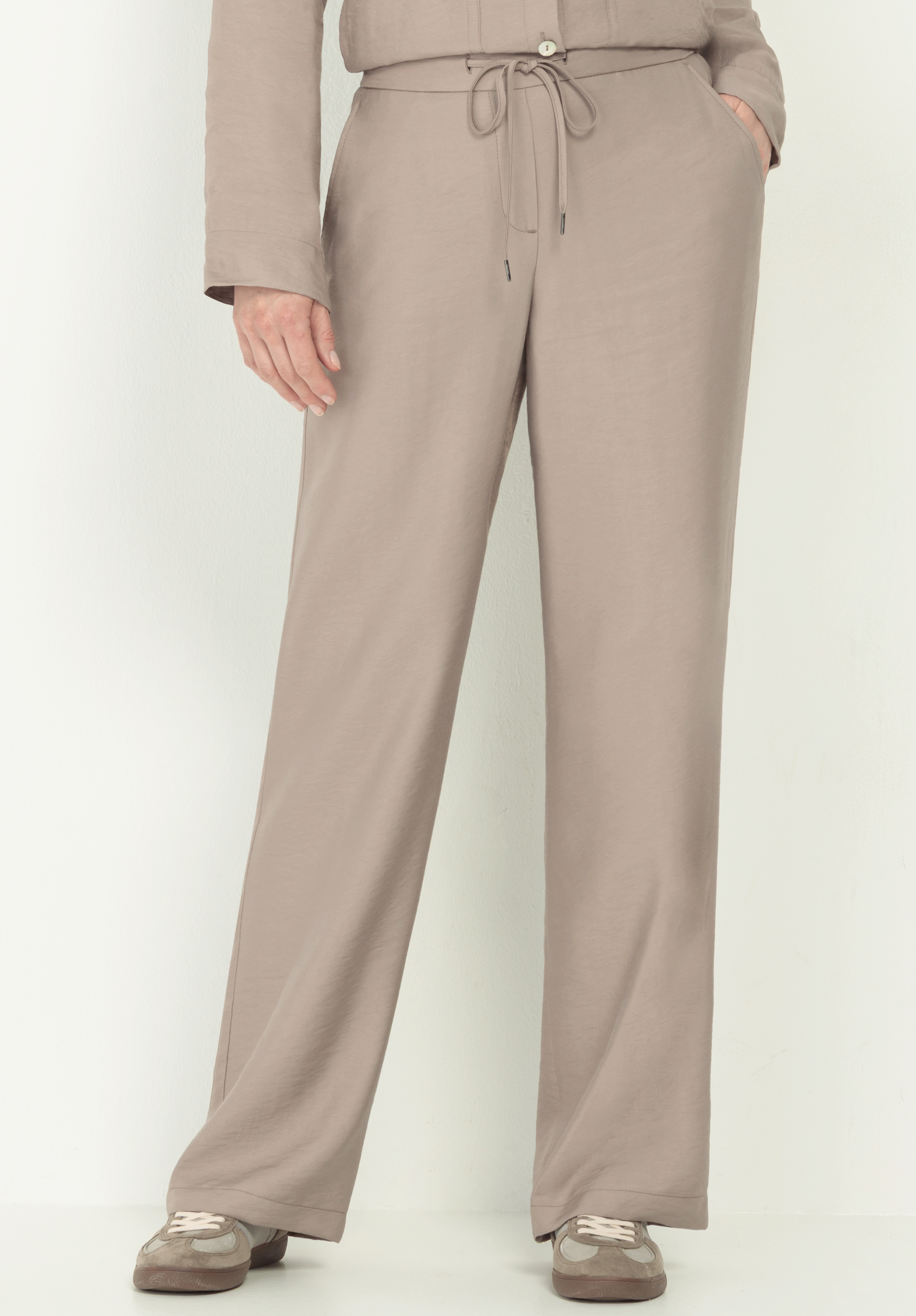 Schlupfhose PARIGI* Person, die eine lockere taupefarbene Hose mit Kordelzug in der Taille und Taschen trägt, gepaart mit taupefarbenen und weißen Turnschuhen. Nur der untere Teil des Oberkörpers und die Beine sind vor einem schlichten hellen Hintergrund zu sehen.