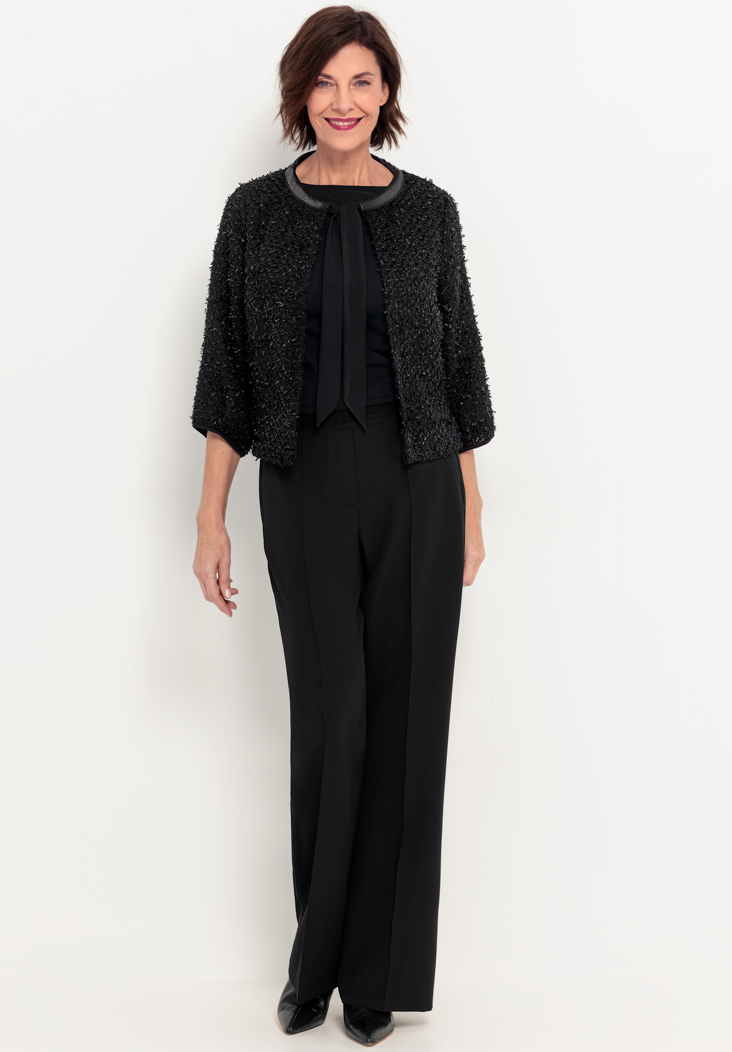 Eine Frau mit kurzen braunen Haaren steht vor einem schlichten weißen Hintergrund und lächelt. Sie trägt eine strukturierte schwarze Jacke, eine schwarze Bluse mit Schleife, eine schwarze Hose mit weitem Bein und schwarze Schuhe.