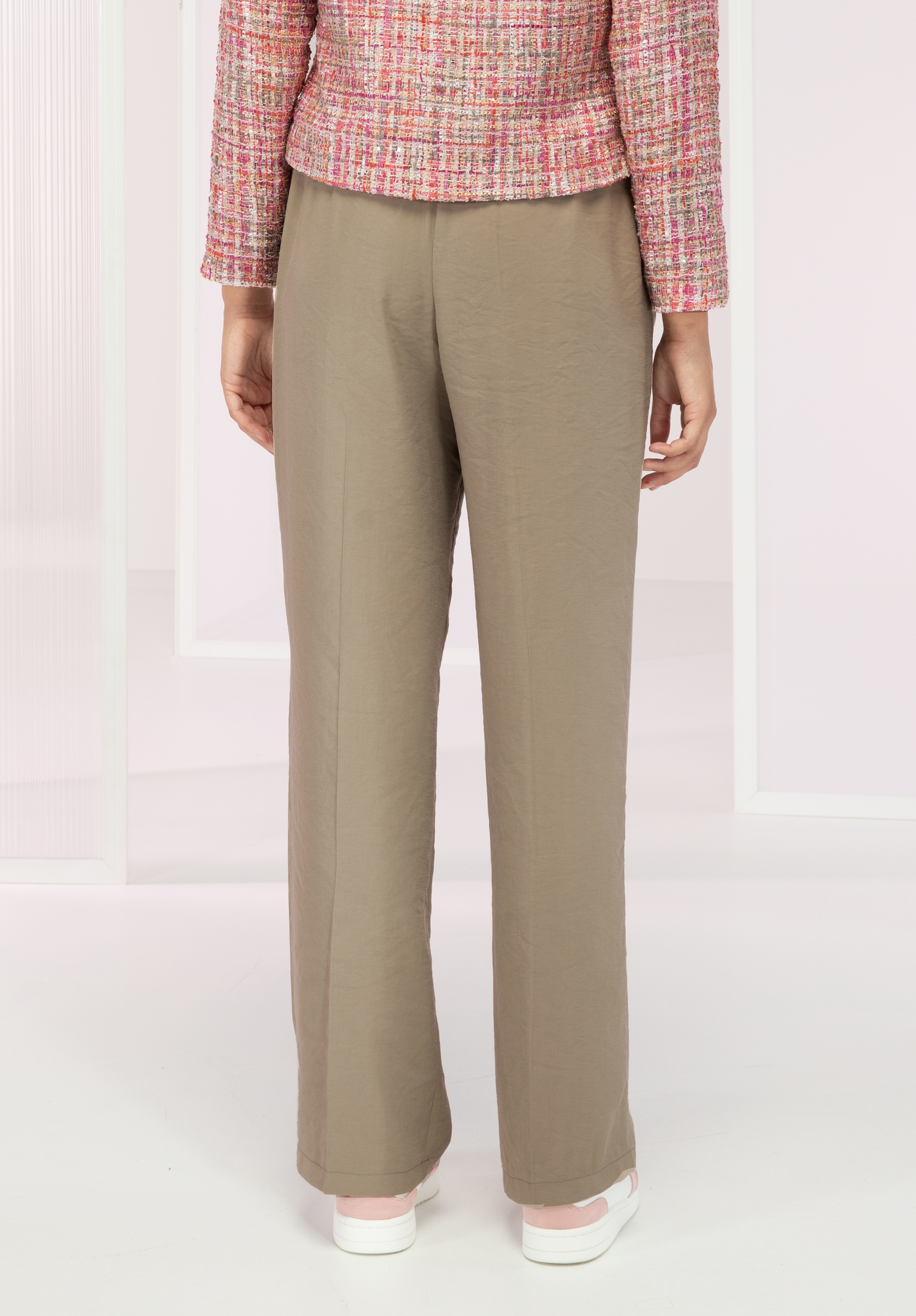 Eine Person steht mit abgewandtem Gesicht da und trägt eine taupefarbene Hose mit geradem Bein, eine rosa-weiß strukturierte Jacke und weiße Schuhe vor einem minimalistischen hellen Hintergrund mit weißen rechteckigen Rahmen.