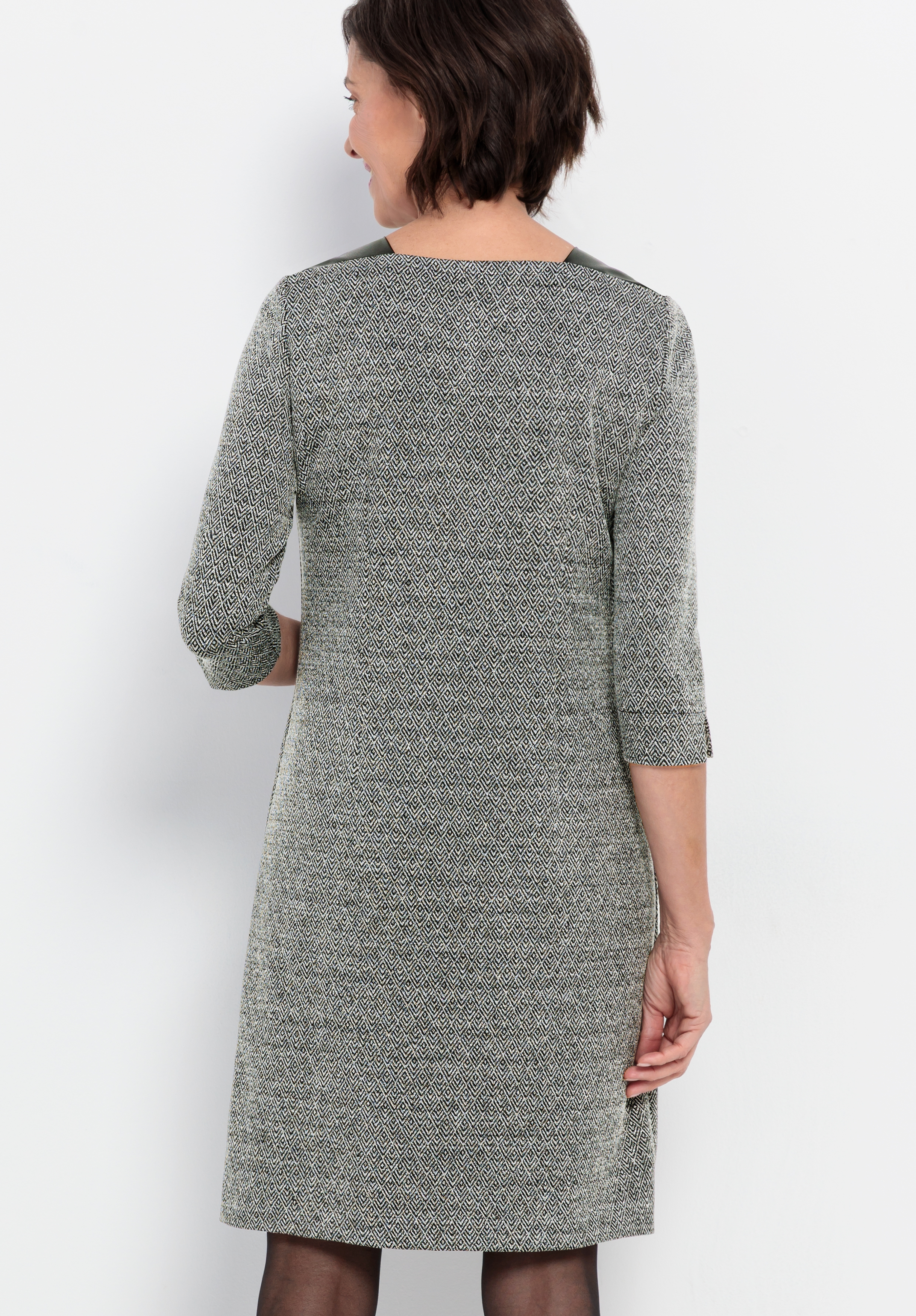 Eine Frau mit kurzen braunen Haaren ist von hinten zu sehen; sie trägt ein knielanges, langärmeliges, grau gemustertes Kleid. Sie steht vor einem schlichten weißen Hintergrund.