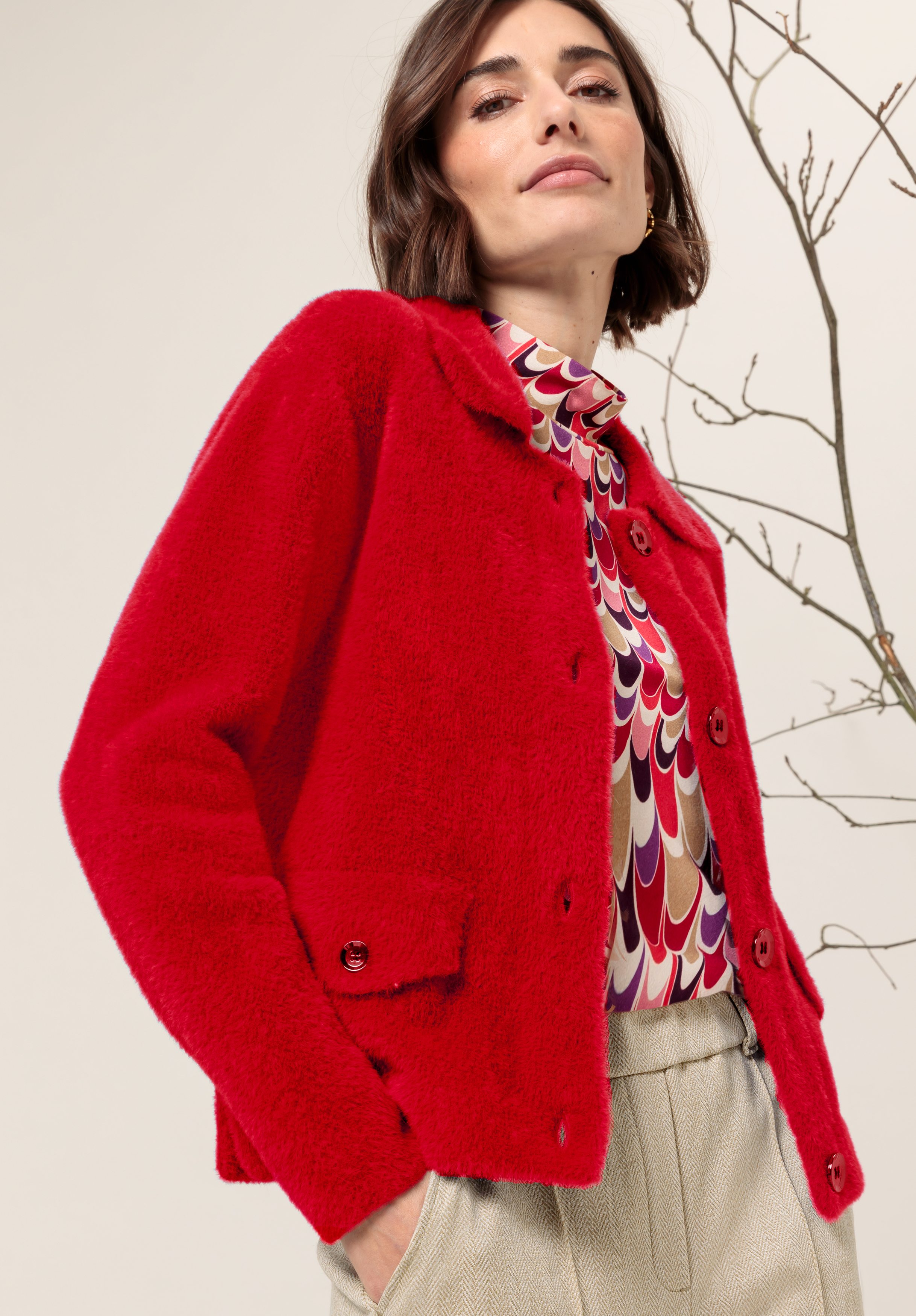 Eine Frau, die eine leuchtend rote Jacke mit Knöpfen über einer bunt gemusterten Bluse und hellbeigen Hosen trägt, steht mit einer Hand in der Tasche da und posiert selbstbewusst vor einem neutralen Hintergrund mit einem kahlen Ast.