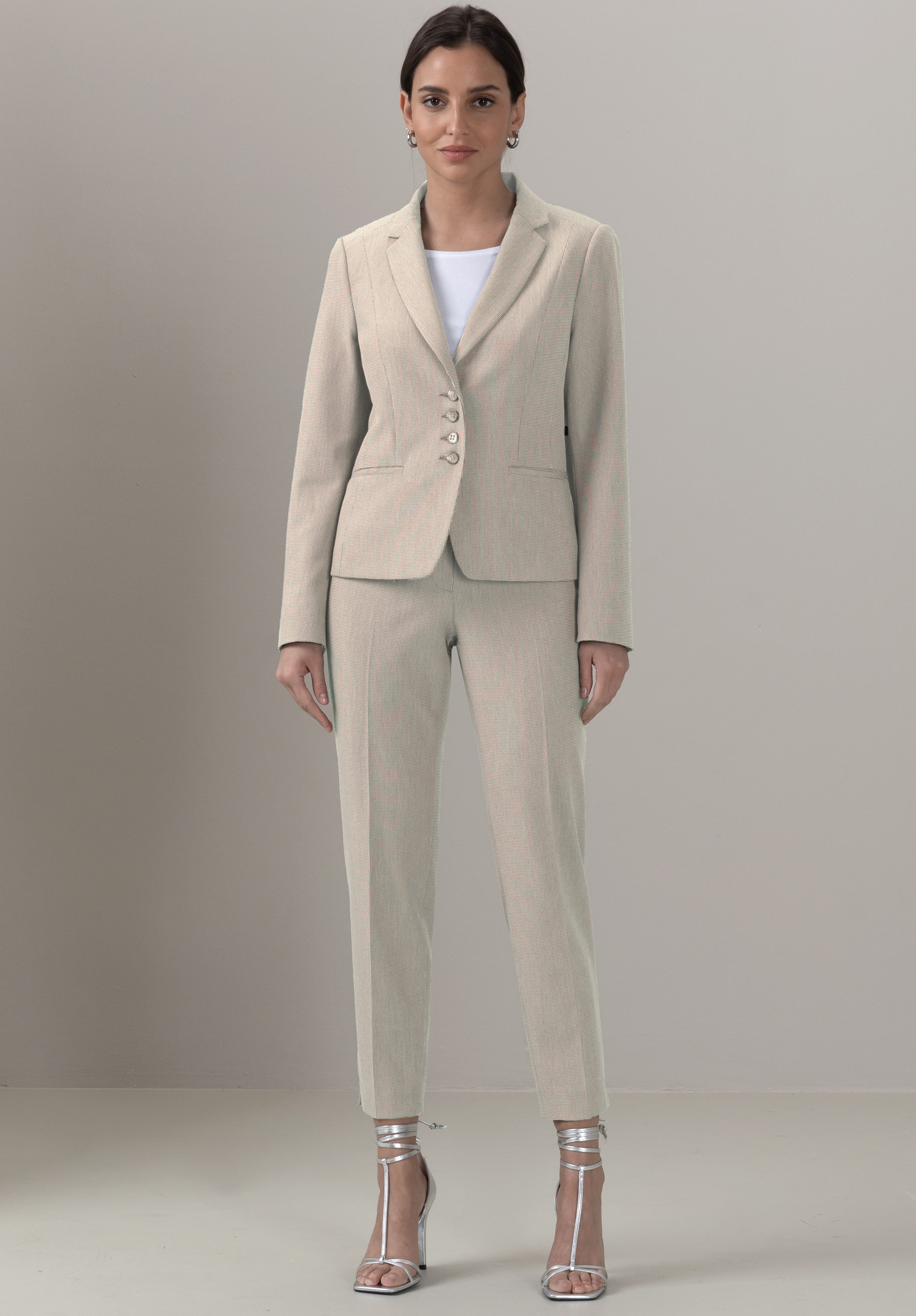 Eine Frau steht in einem cremefarbenen Hosenanzug mit weißer Bluse und silbernen hochhackigen Sandalen vor einem schlichten hellgrauen Hintergrund. Sie blickt mit neutraler Miene nach vorne.