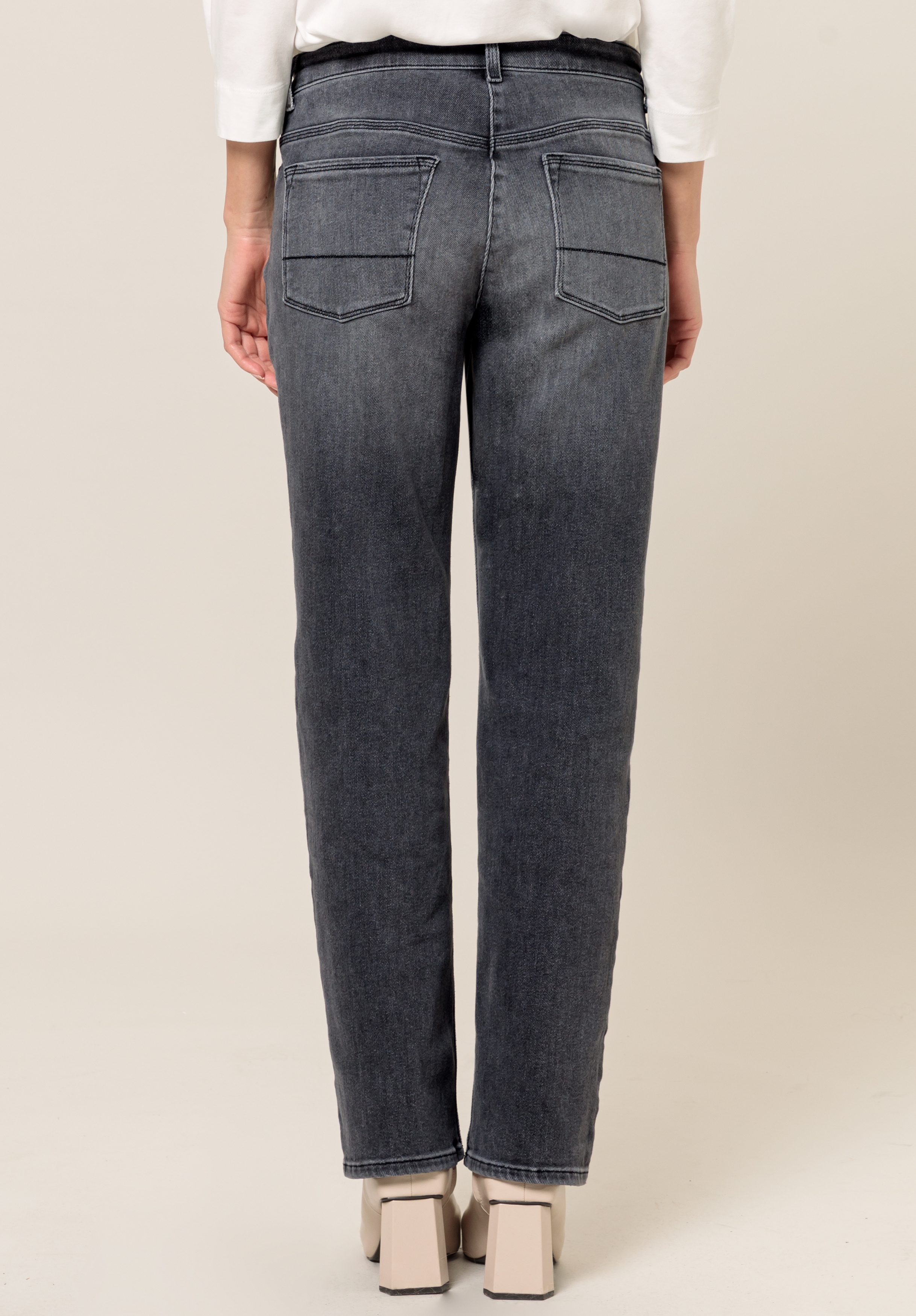 Eine Person trägt gerade geschnittene, dunkelgraue Jeans mit Gesäßtaschen und cremefarbene Schuhe mit Absatz, von hinten vor einem schlichten hellen Hintergrund gezeigt.