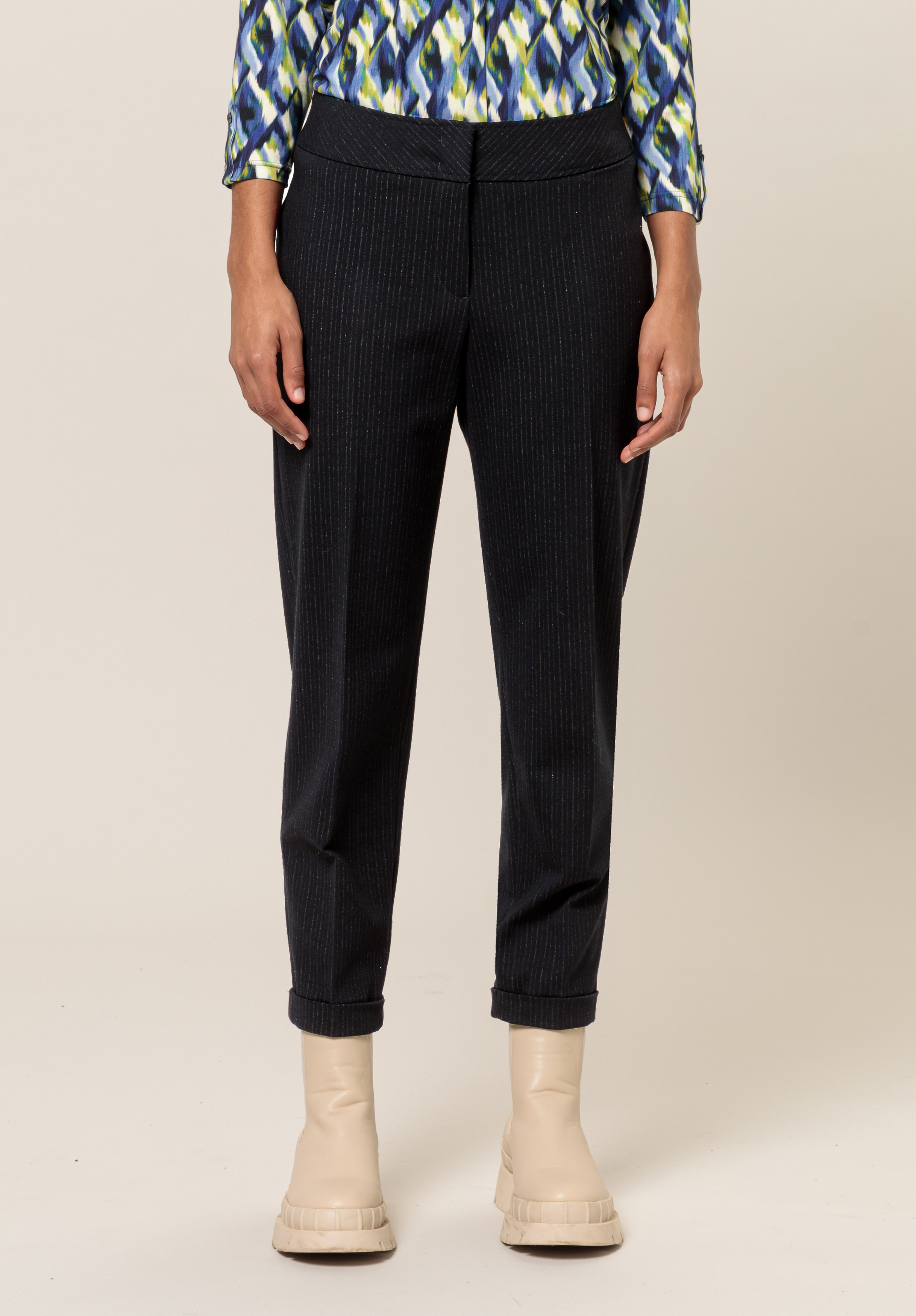 Eine Person in einer dunklen Nadelstreifenhose, einem gemusterten blau-grünen Oberteil und beigen Stiefeletten steht vor einem schlichten beigen Hintergrund und ist von den Schultern bis zu den Füßen sichtbar.