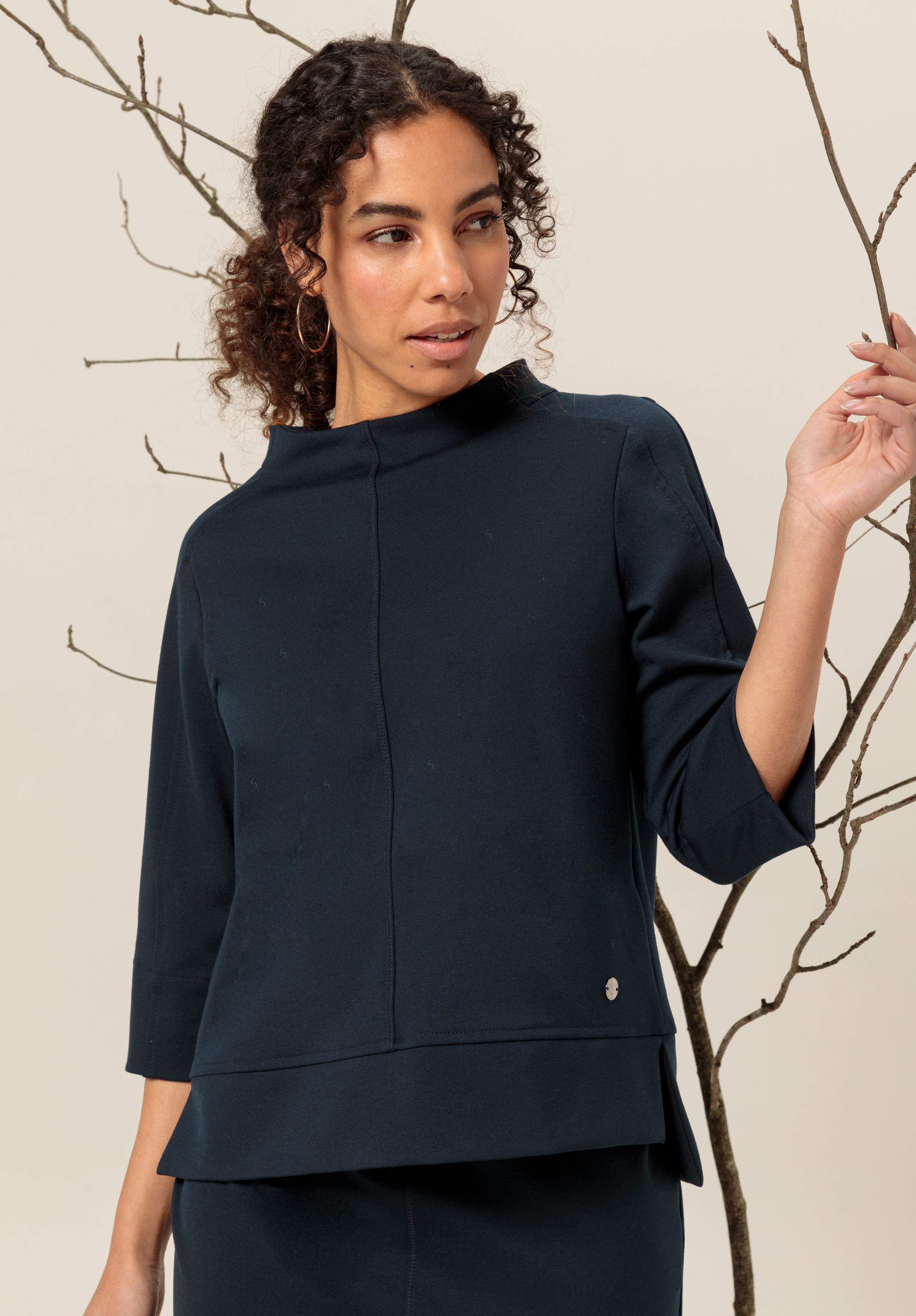 3/4-Arm-Shirt IDA Eine Frau mit lockigem Haar und einem dunklen Langarmshirt steht vor einem beigen Hintergrund mit dünnen, blattlosen Zweigen. Sie blickt zur Seite und hat eine Hand in die Nähe der Zweige erhoben.