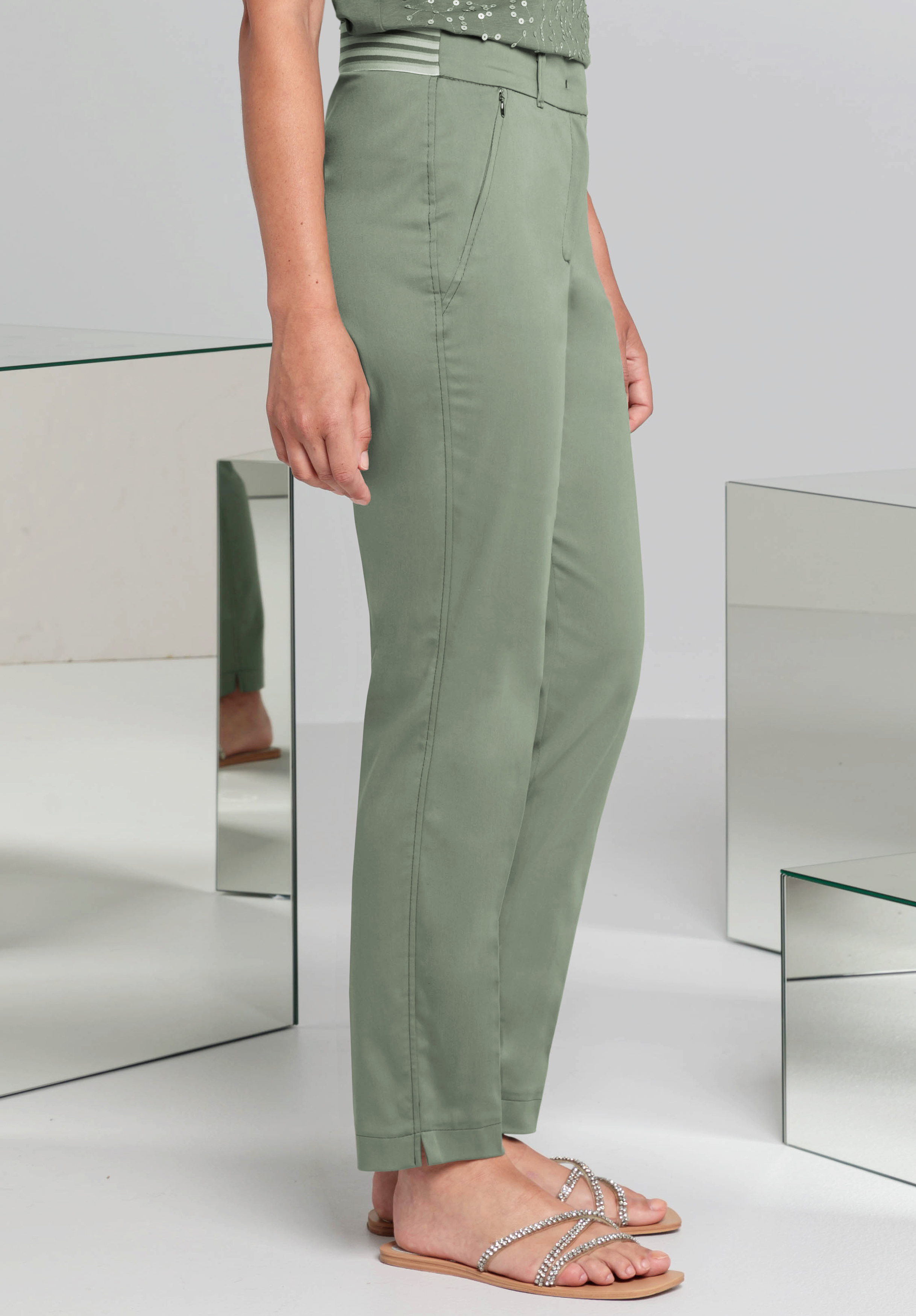 Eine Person steht seitlich und trägt eine salbeigrüne Hose und verzierte Sandalen. Im Hintergrund sind Spiegel und eine helle, minimalistische Atmosphäre zu sehen.