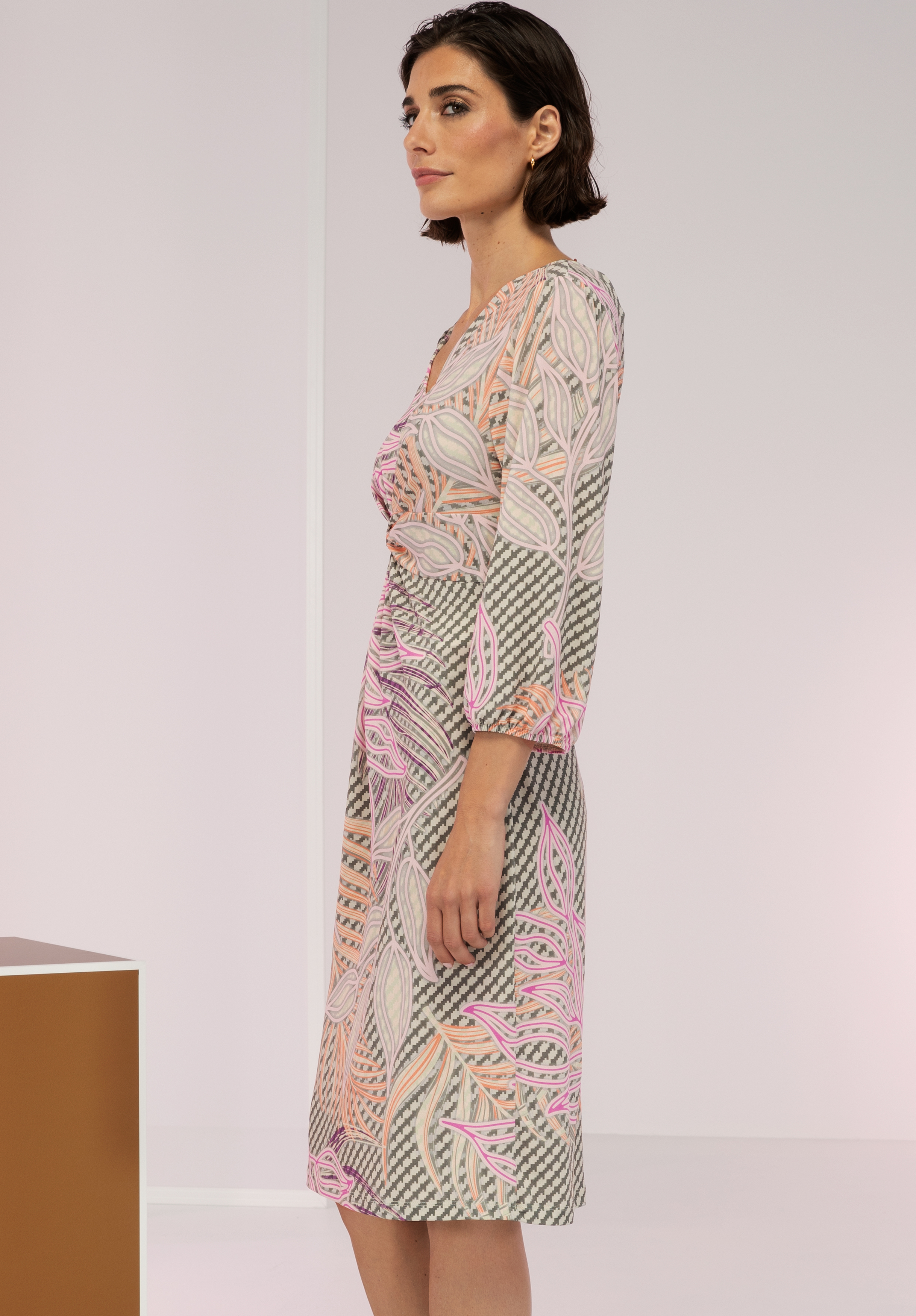 Eine Frau mit kurzen dunklen Haaren steht im Profil und trägt ein knielanges Kleid mit einem abstrakten Blattmuster in Rosa, Grau und Beige. Der Hintergrund ist minimalistisch und sanft beleuchtet.