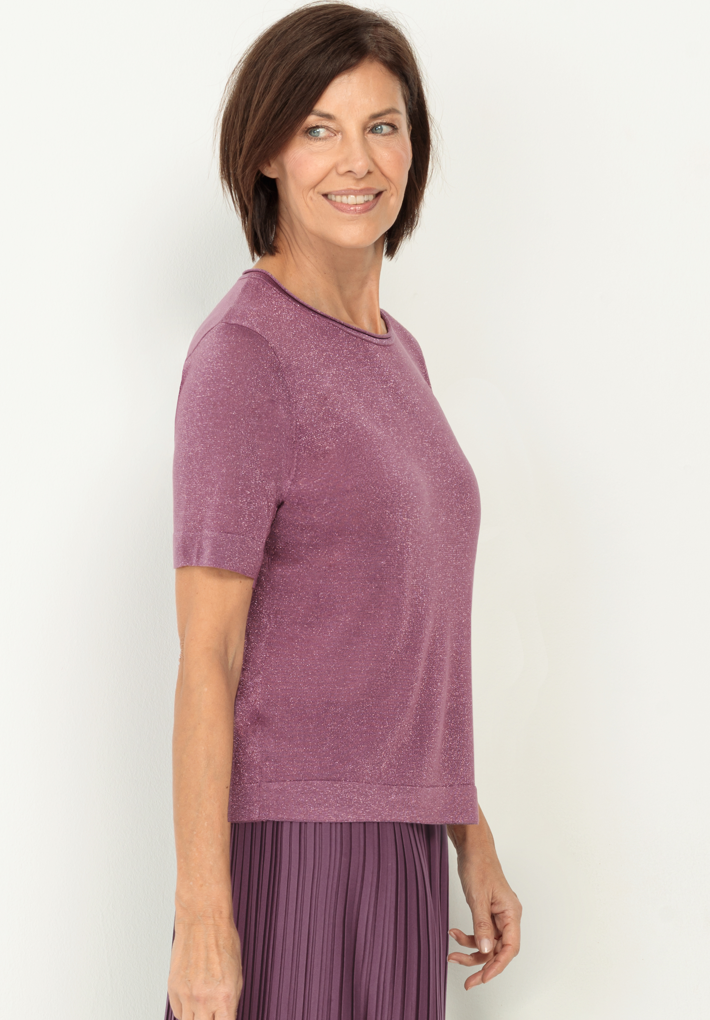 Eine Frau mit kurzen braunen Haaren, die ein kurzärmeliges, lila schimmerndes Oberteil und einen passenden Faltenrock trägt, steht lächelnd im Profil vor einem schlichten weißen Hintergrund.