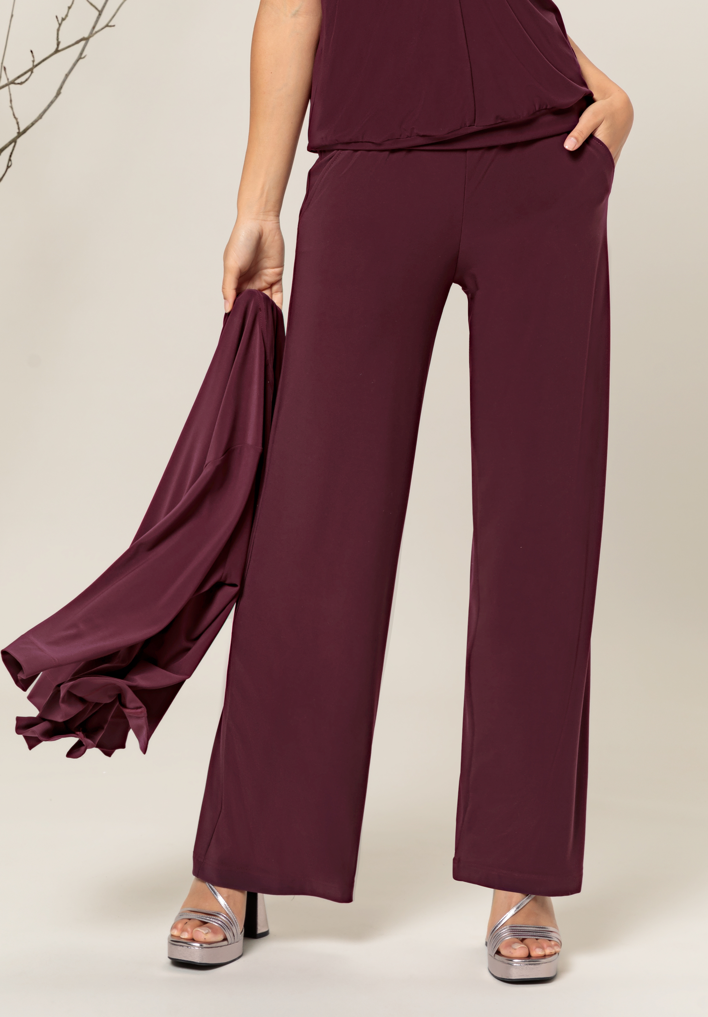 Eine Person in hoch taillierten, weiten kastanienbraunen Hosen und silbernen Plateauschuhen steht mit einer Hand in der Tasche da und hält vor einem neutralen Hintergrund ein passendes kastanienbraunes Kleidungsstück in der anderen Hand.