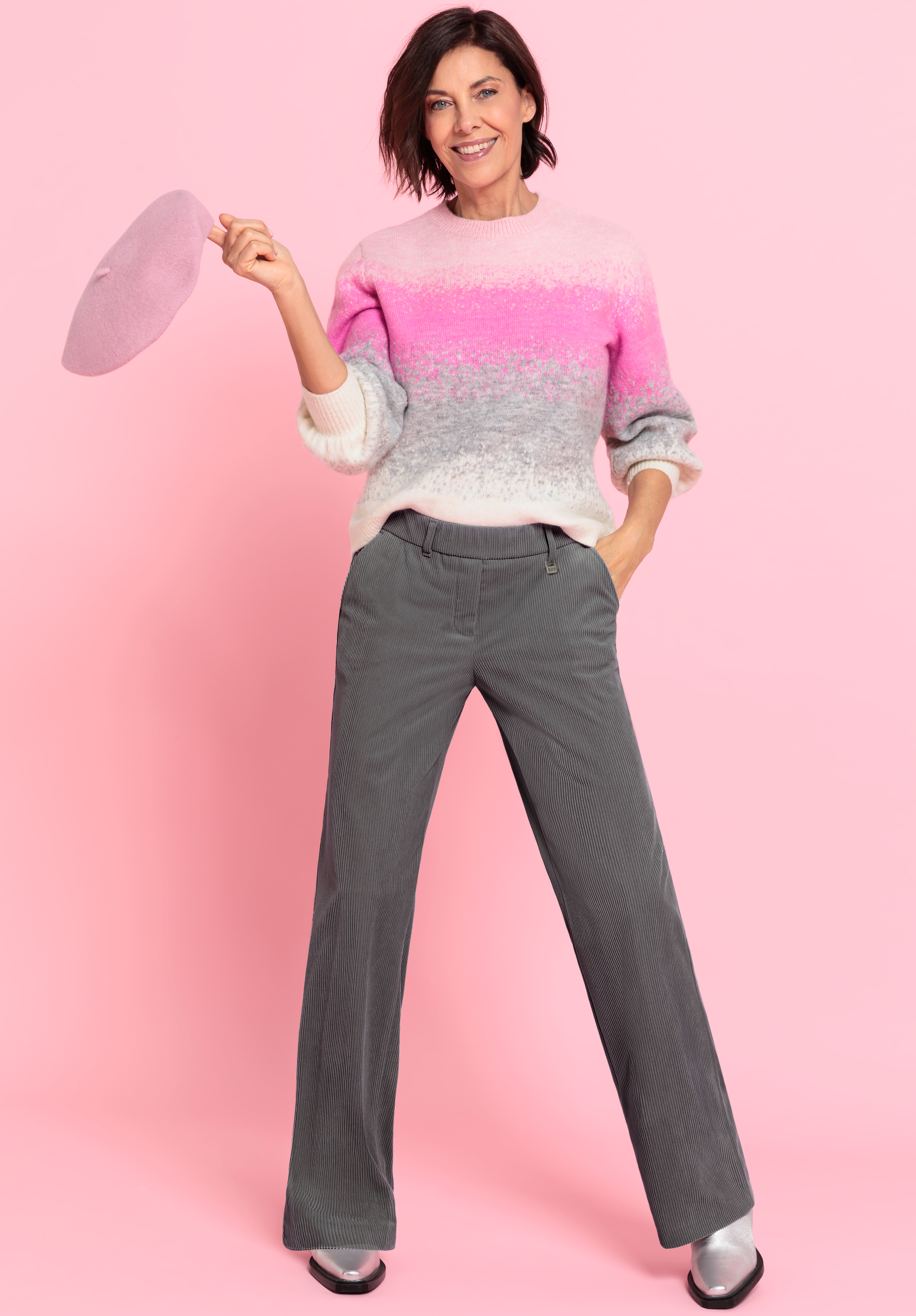 Eine Frau steht lächelnd vor einem rosafarbenen Hintergrund, trägt einen rosa-weißen Ombre-Pullover, eine graue Hose und silberne Schuhe und hält in einer Hand eine rosa Baskenmütze.