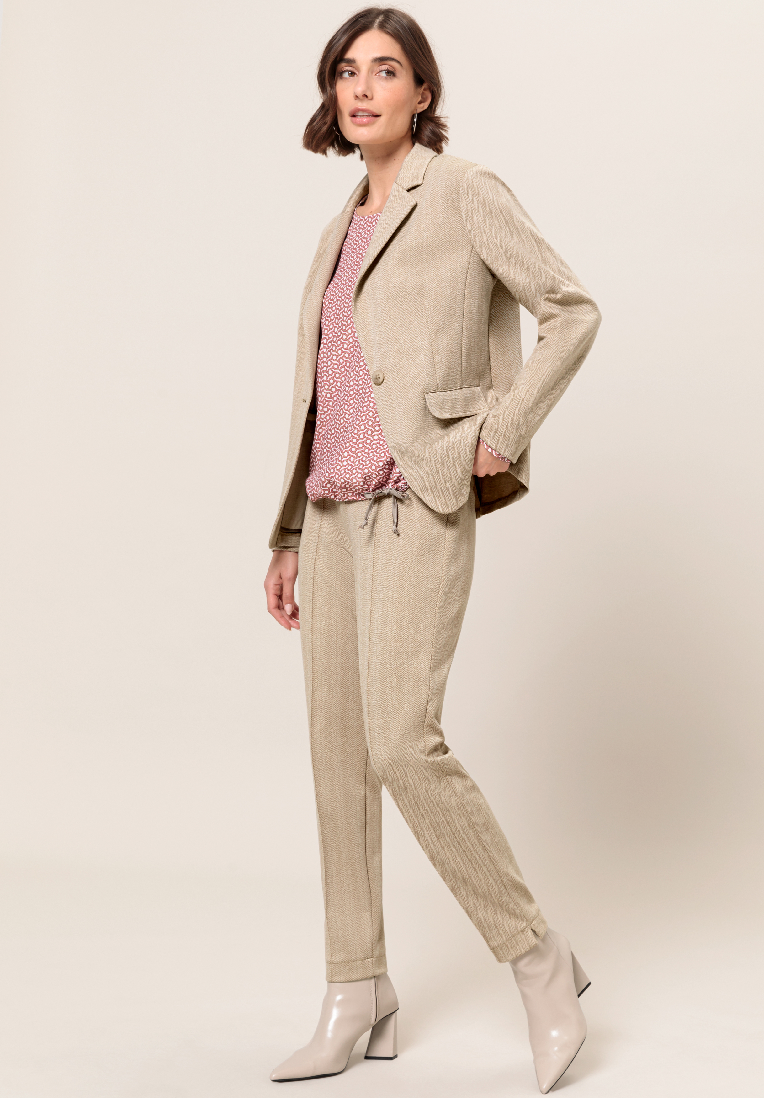 Eine Frau mit kurzen braunen Haaren trägt einen hellbeigen Hosenanzug, eine gemusterte rot-weiße Bluse und hellbeige Stiefeletten. Sie steht vor einem schlichten Hintergrund und posiert mit einer Hand in der Tasche.