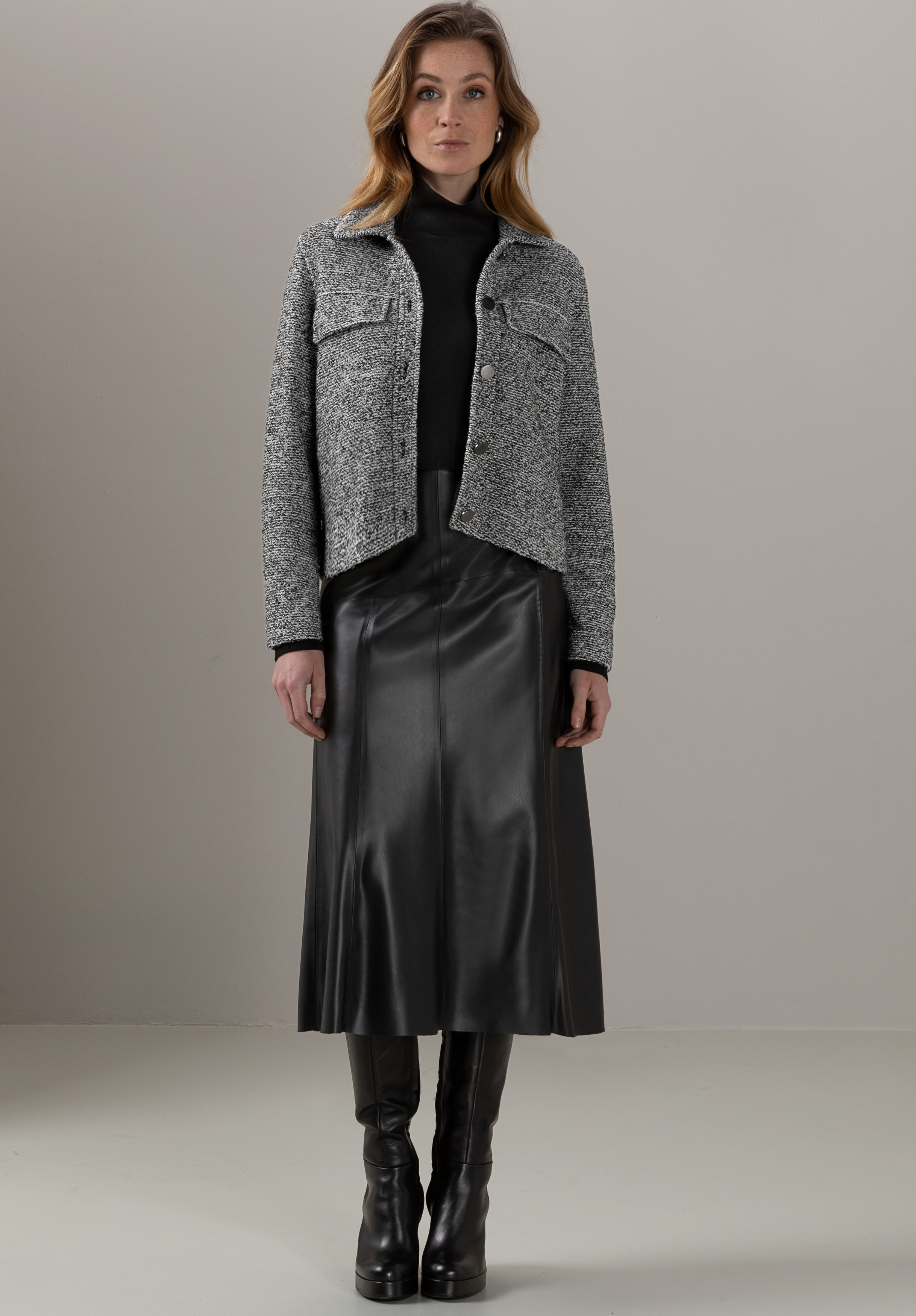 Eine Frau steht mit Blick nach vorne vor einem schlichten, hellen Hintergrund und trägt eine graue Strukturjacke über einem schwarzen Rollkragenpullover, einen schwarzen Midirock aus Leder und schwarze kniehohe Stiefel.